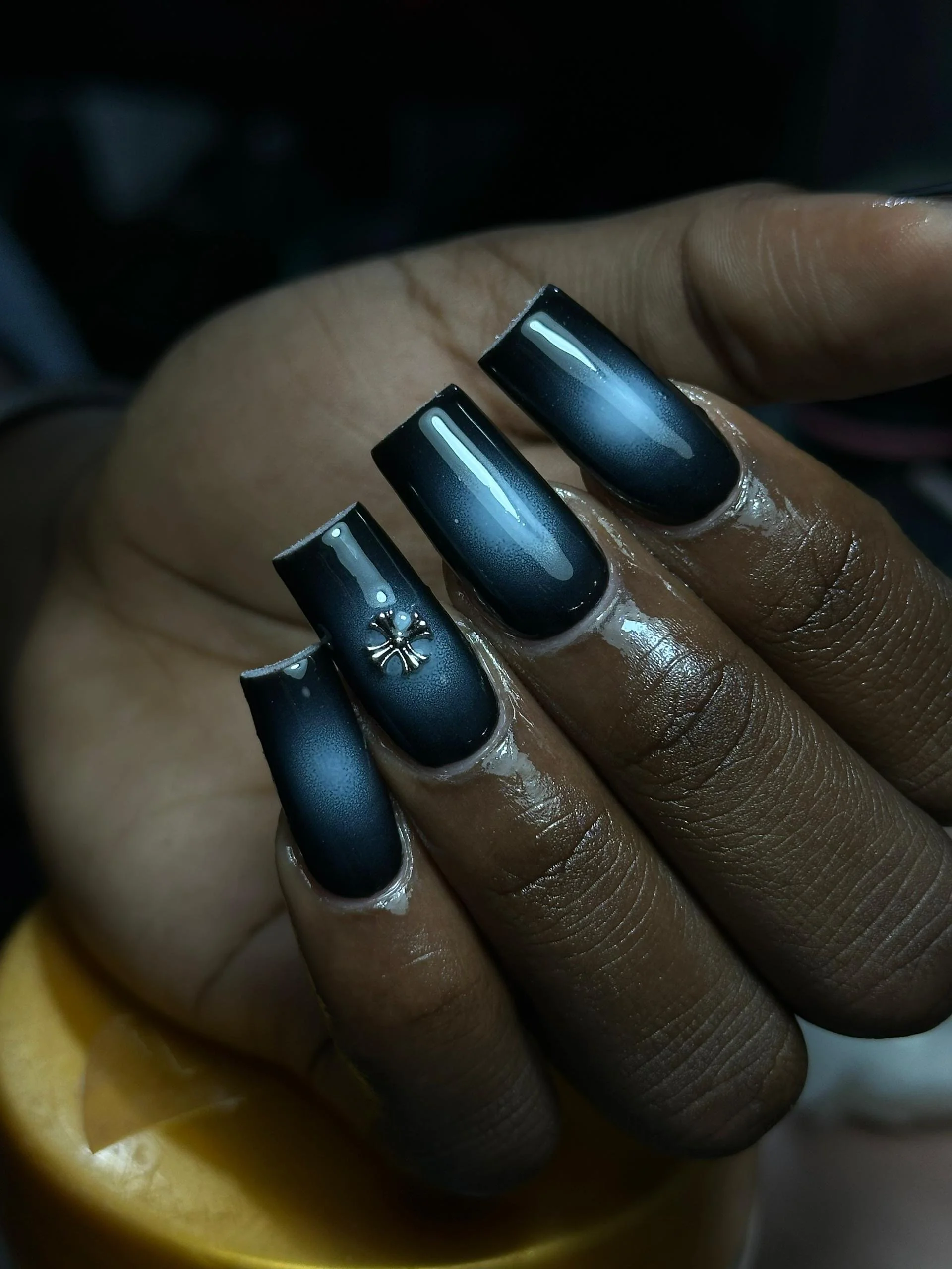 Close-up of a hand with dark, glossy, ombre black to teal nails and a small silver decorative charm on the ring finger.