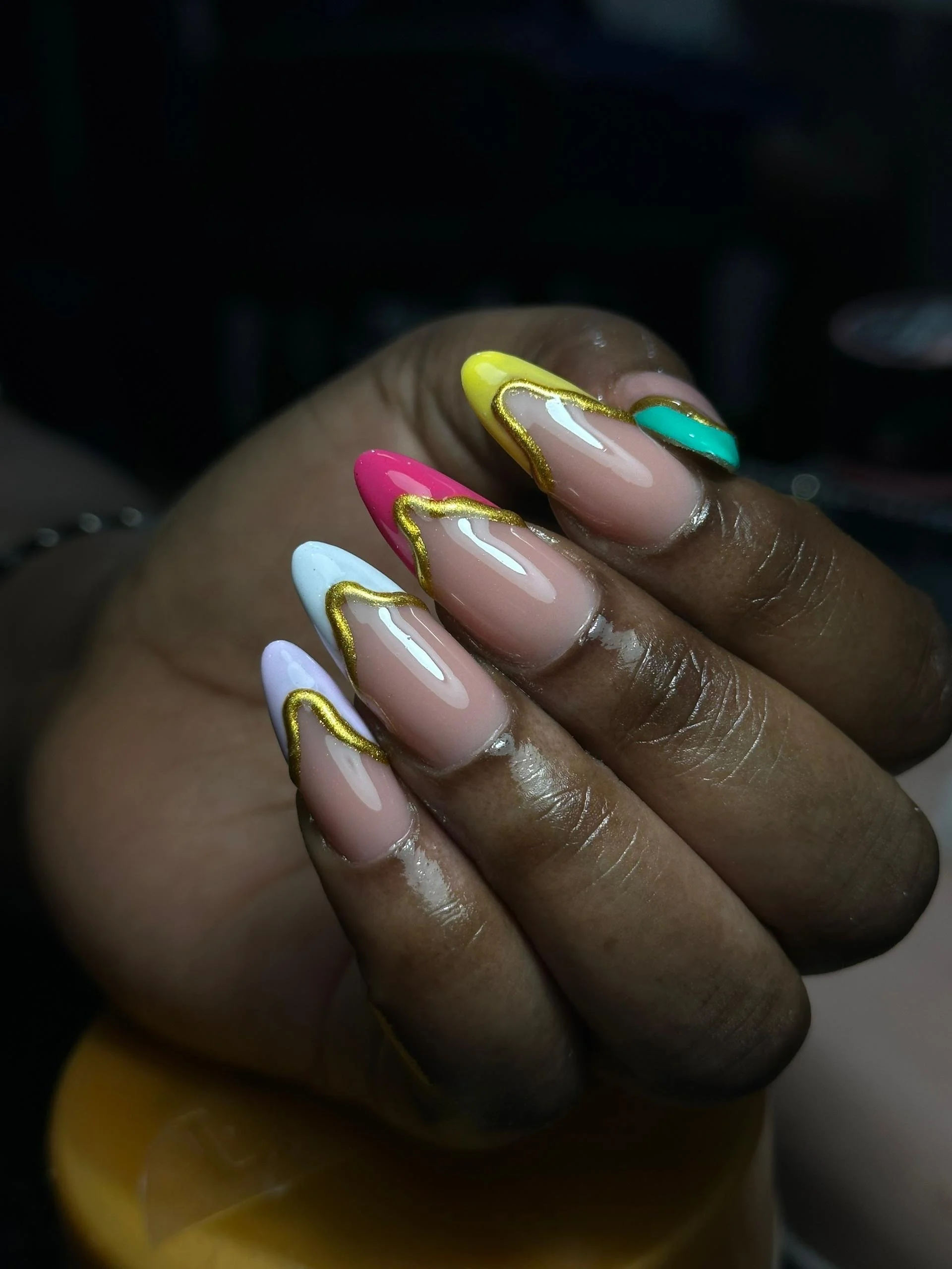 Manicured fingernails with pink, white, yellow, and teal nail polish, each outlined with gold accents.