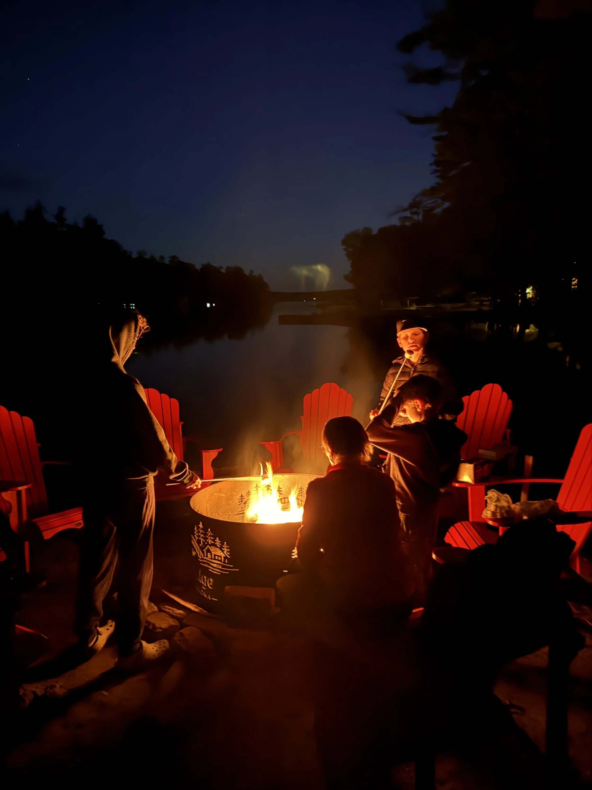 Group of people gathered outdoors around a campfire at night, with trees and a body of water in the background.