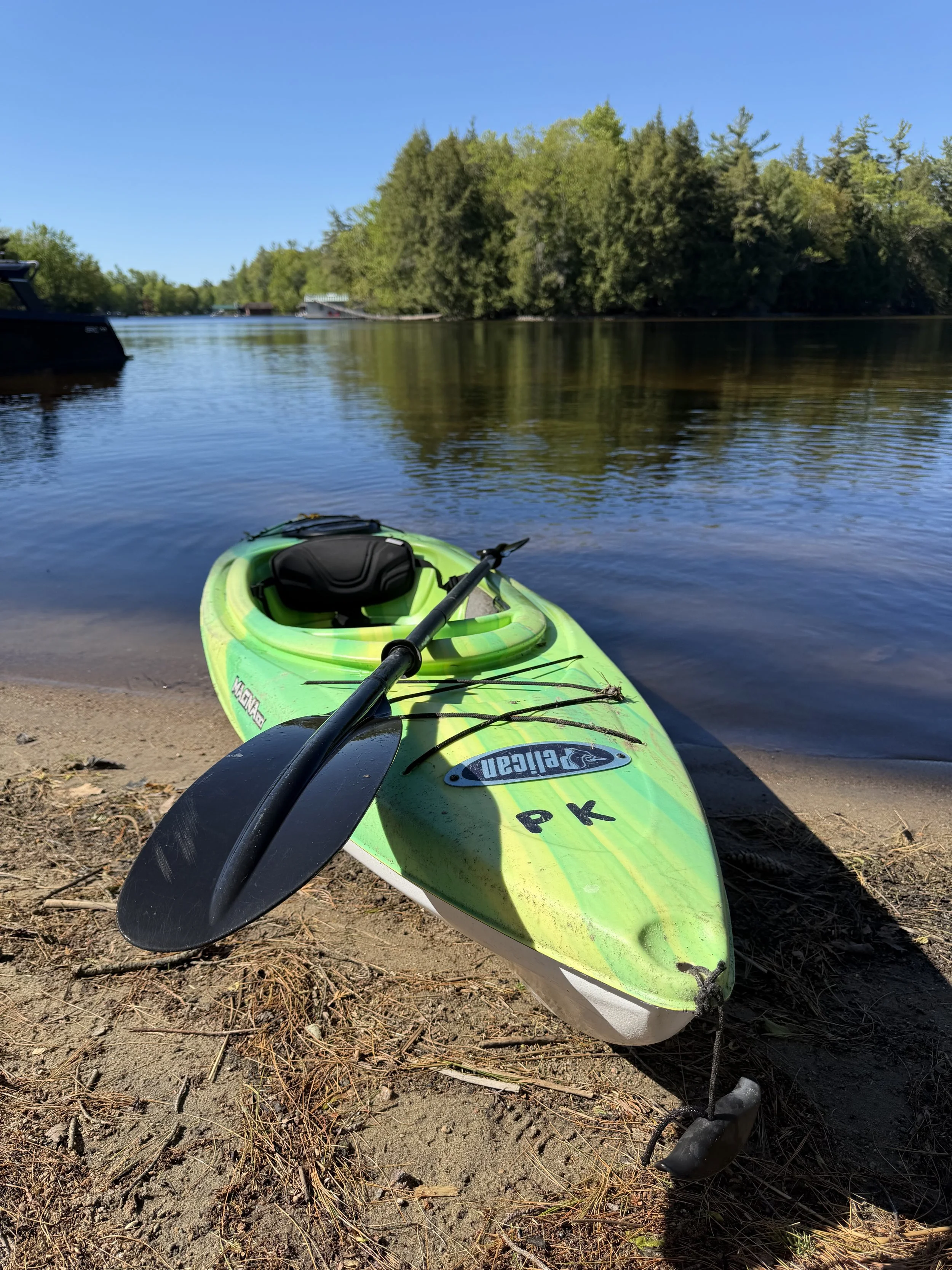 Green kayak on sandy shore next to calm river, surrounded by trees under blue sky.
