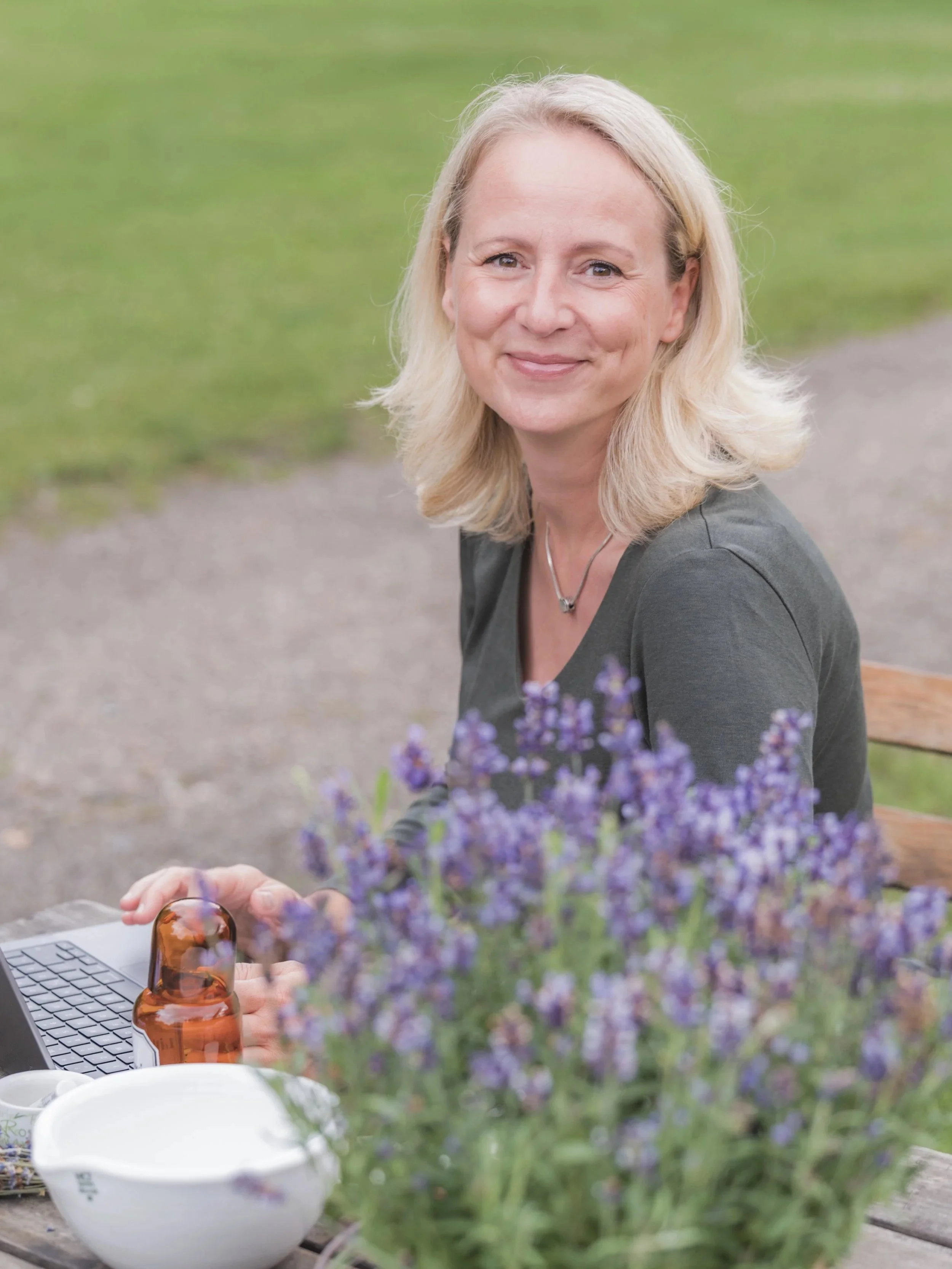 Eine blonde Frau sitzt im Freien vor einem grünen Hintergrund, lächelt in die Kamera, während sie mit einem Laptop arbeitet. Im Vordergrund sind violette Lavendelblumen und eine braune Flasche sichtbar.