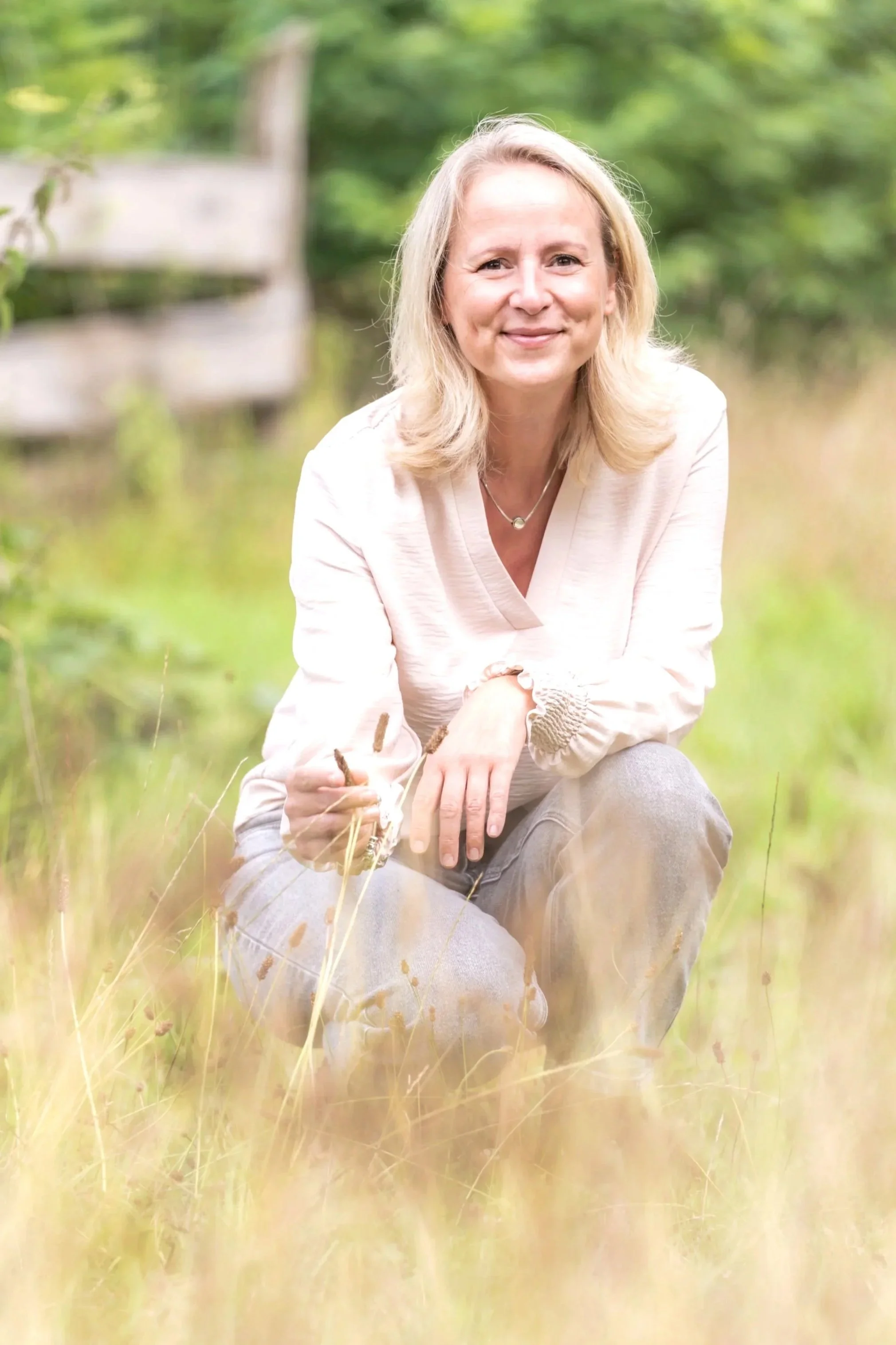 Eine Frau sitzt im Gras in einer natürlichen Umgebung mit grünen Bäumen im Hintergrund. Sie lächelt, trägt eine hellfarbene Bluse und eine Halskette.
