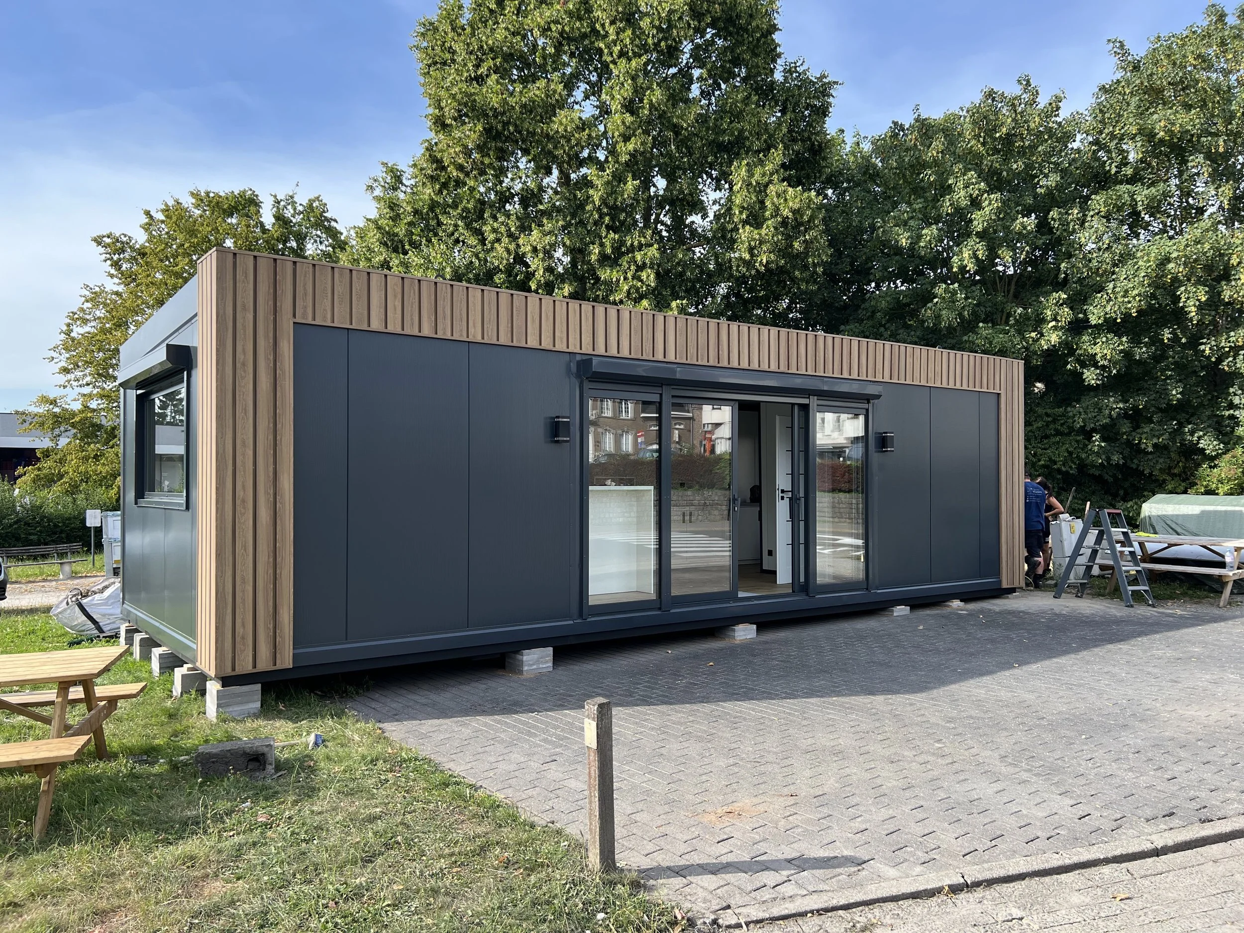 Moderne tiny house met grote ramen, houten en zwarte buitenwanden, geplaatst op betonnen blokken, met een natuurlijke omgeving en bomen op de achtergrond.