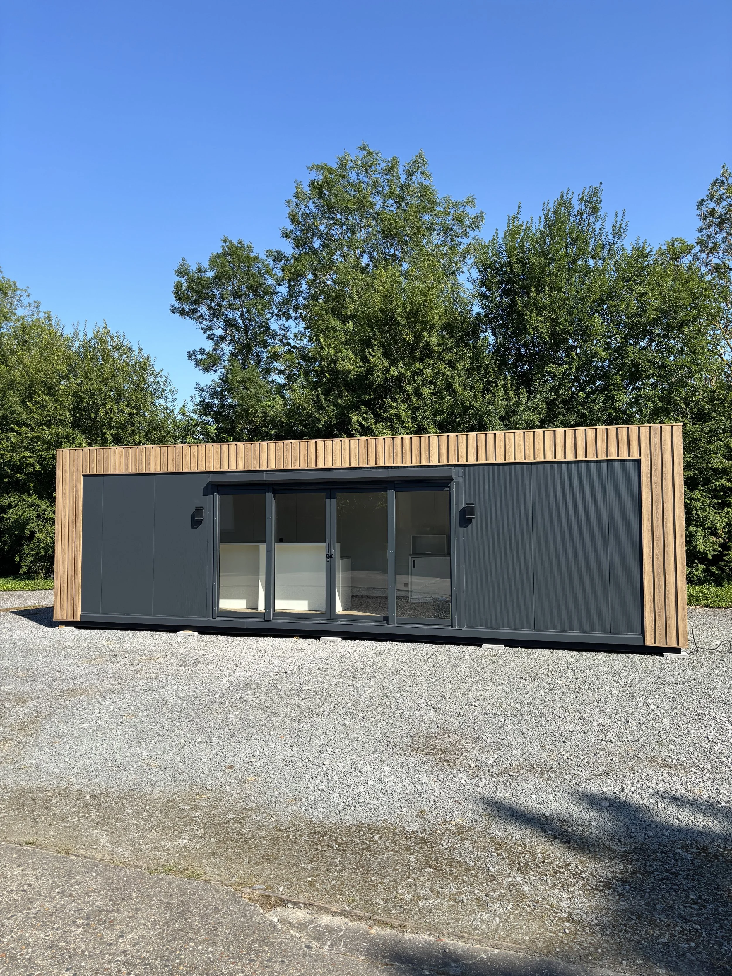 Een modern tiny house met zwarte en houten gevel, grote glazen schuifdeuren, omgeven door groene bomen onder een blauwe hemel.