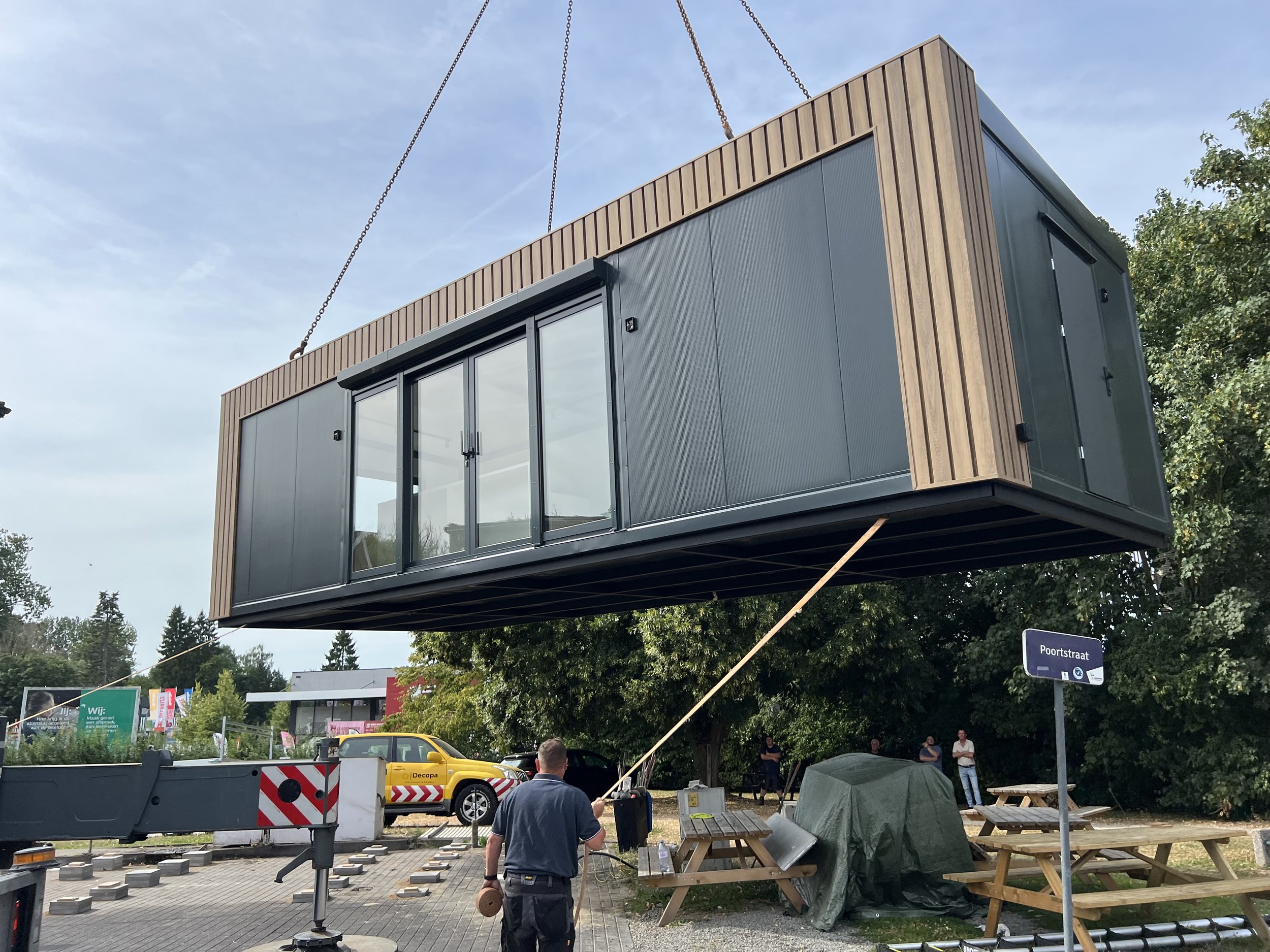 Een overzicht van een parkeerterrein en een groot gebouw met een plat dak, enkele vrachtwagens en auto's geparkeerd, en tijdelijke overkappingen voor goederen of personen.