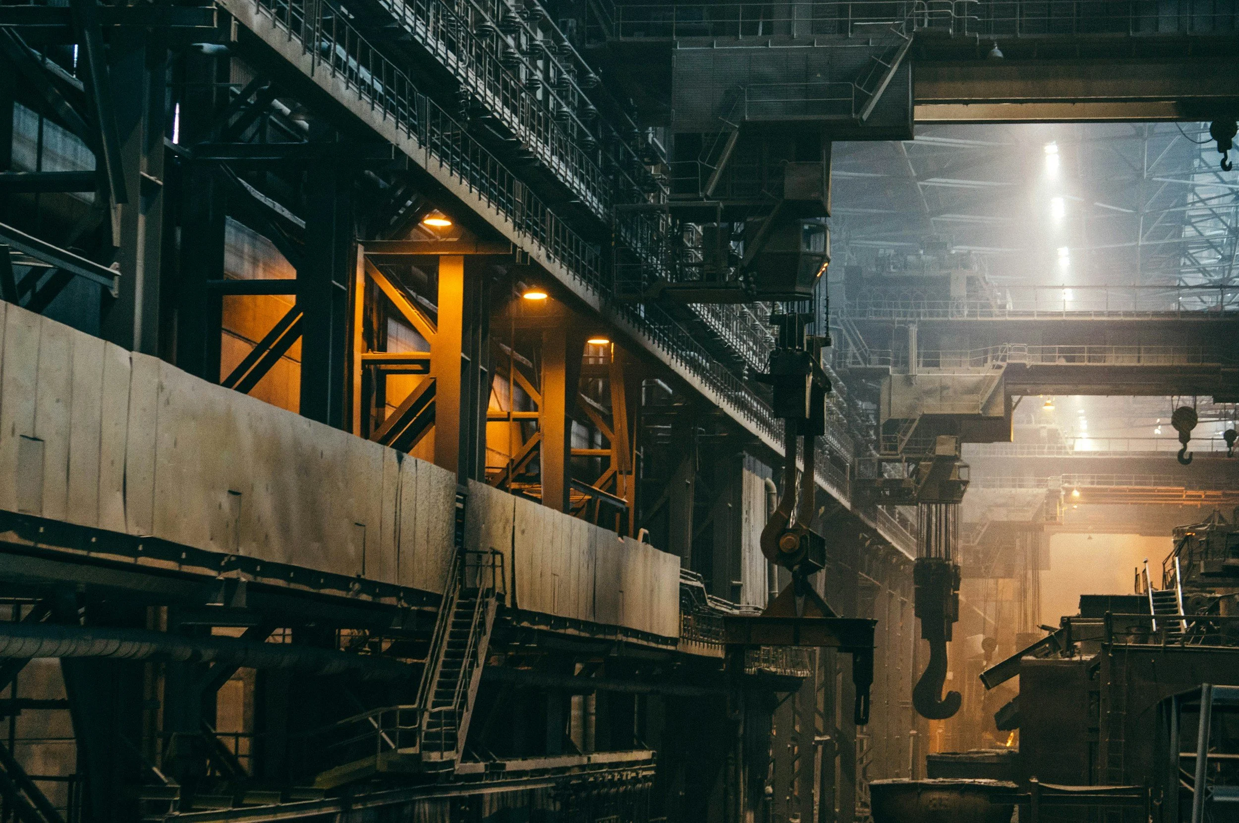 Binnenkant van een grote industriële ruimte met metalen bruggen, trappen en machines, gefilterd door stof en licht.