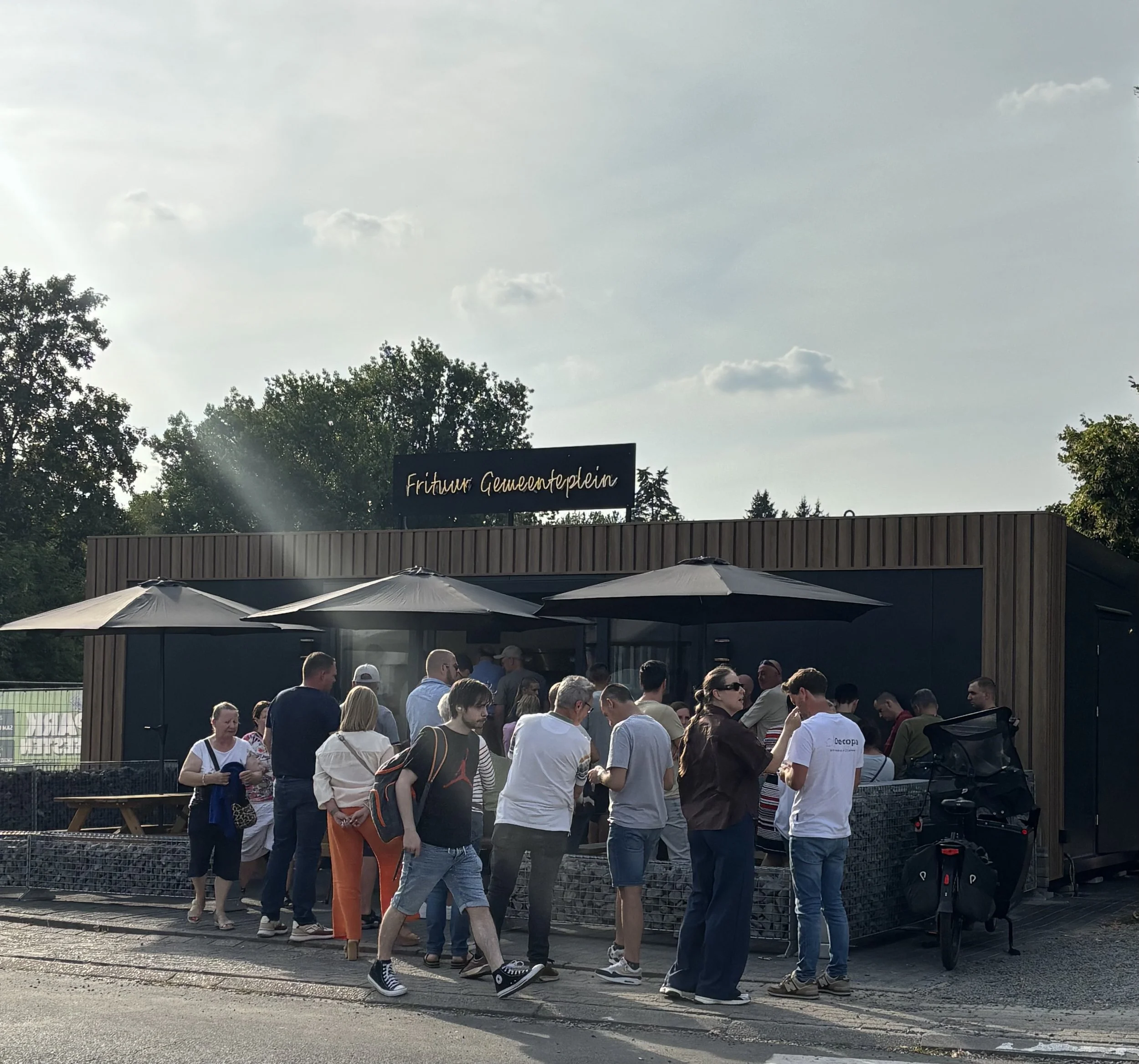 Mensen staan in een wachtrij voor een evenement, met een houten en metalen afscheiding en enkele parasols boven de mensen. Er hangt een bord met de tekst 'Friture Gemeenteplein'.