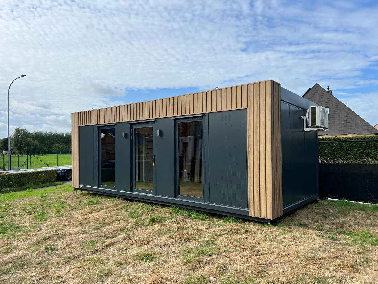 Een graafmachine die een container ophijst in een buitenruimte met gravel en enkele gebouwen op de achtergrond en bomen, onder een bewolkte hemel.