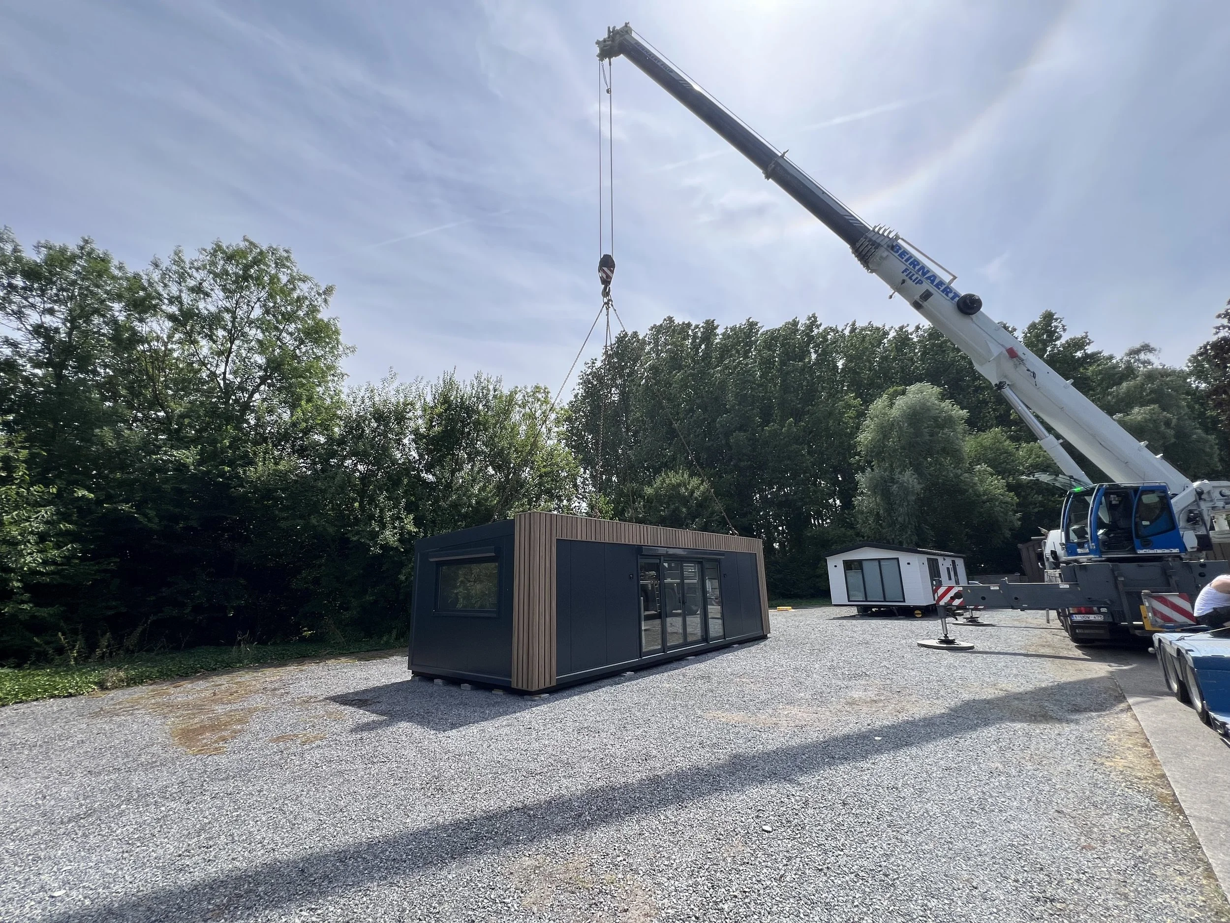 Een graafmachine die een container ophijst in een buitenruimte met gravel en enkele gebouwen op de achtergrond en bomen, onder een bewolkte hemel.