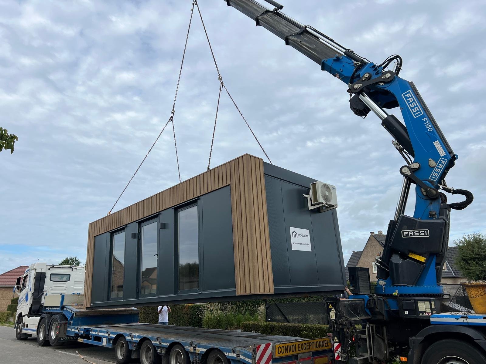 Een modern tiny house met zwarte en houten gevel, grote glazen schuifdeuren, omgeven door groene bomen onder een blauwe hemel.