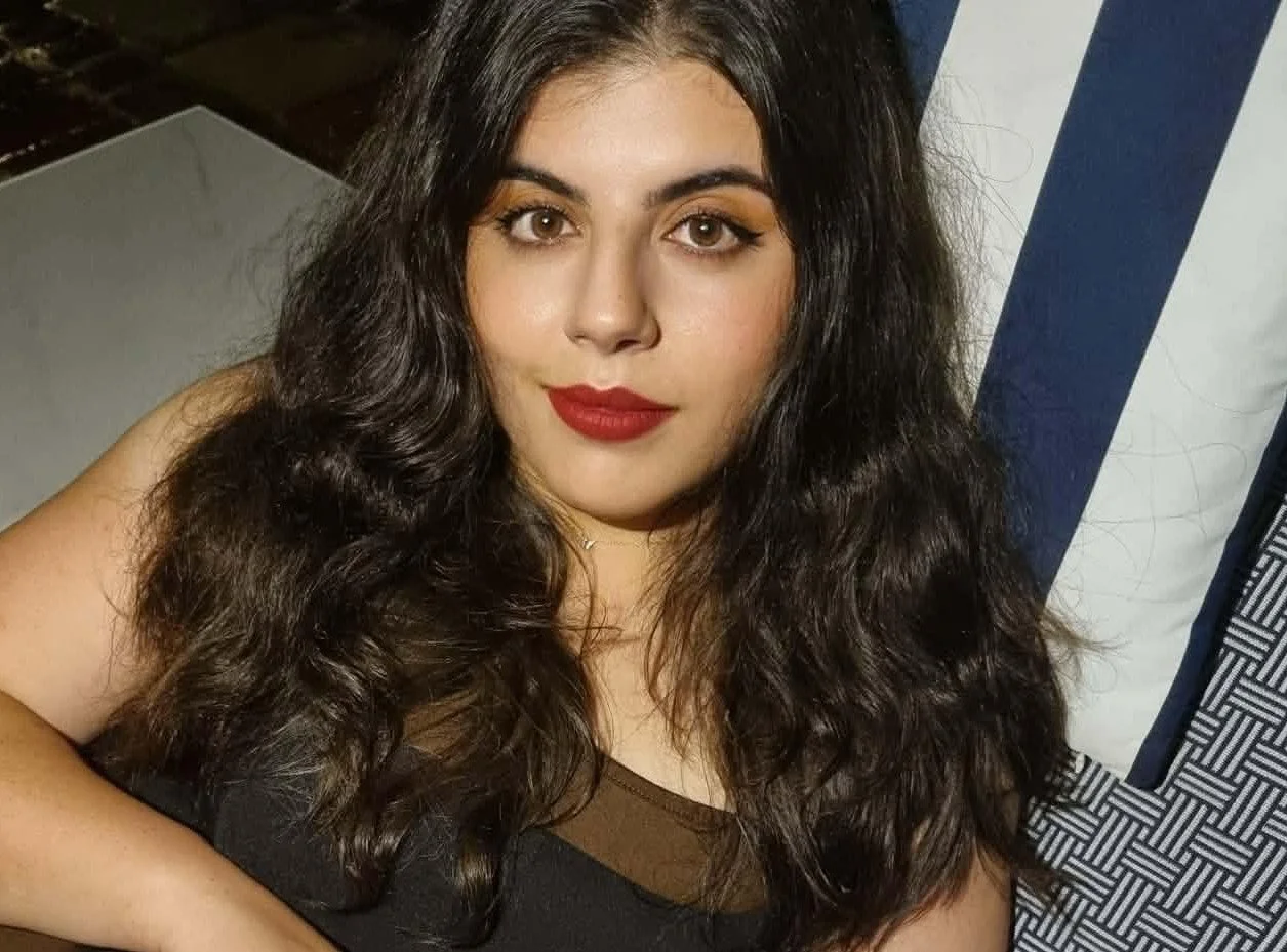 A woman with long, dark, wavy hair and red lipstick looking at the camera