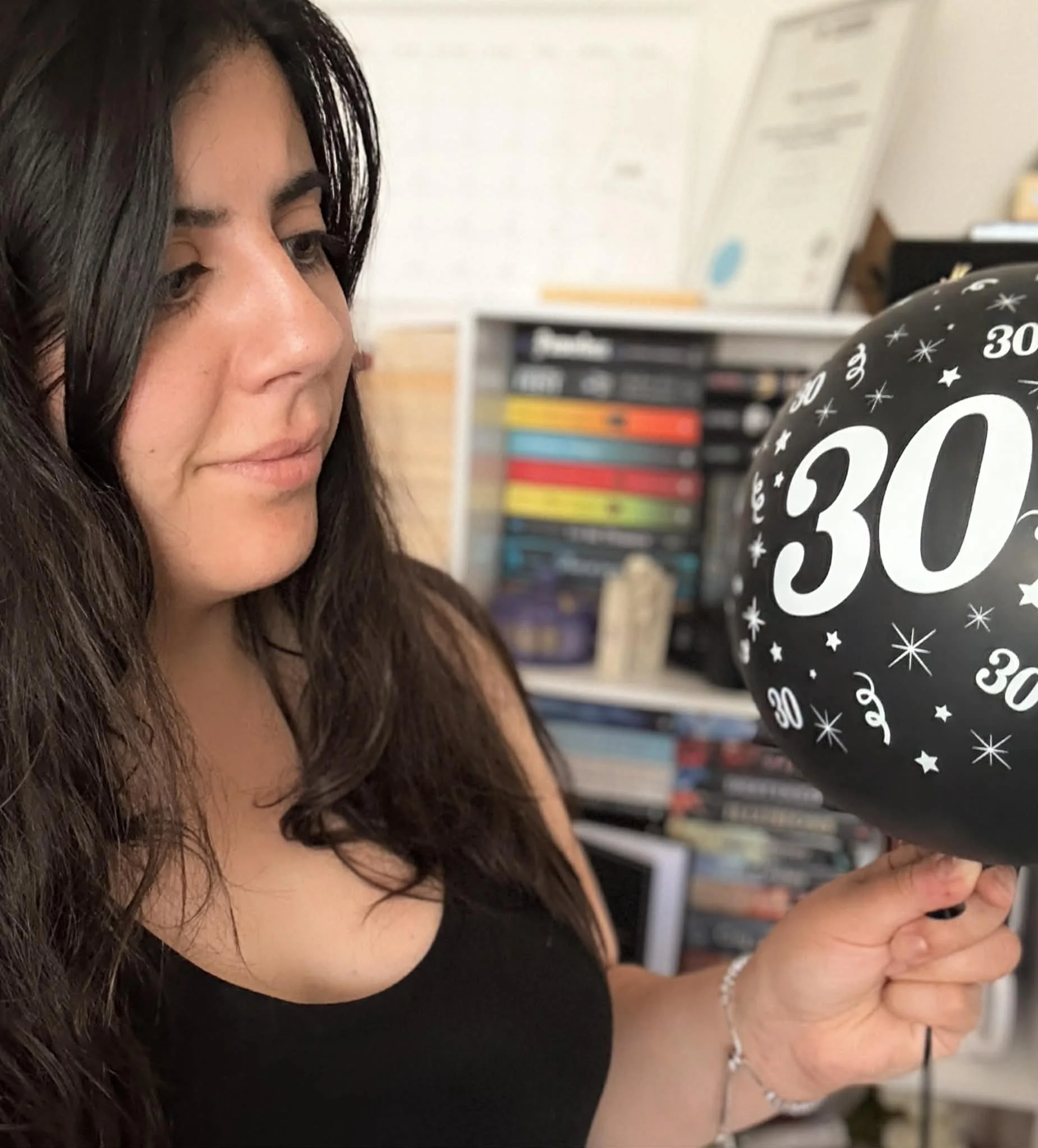 A woman with long dark hair holding a black birthday balloon with white '30' printed on it, in a room with bookshelves in the background.