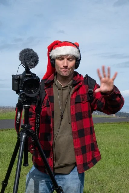 A man wearing a Santa hat, headphones, and a red plaid jacket is standing outdoors with a camera on a tripod, gesturing with his right hand.