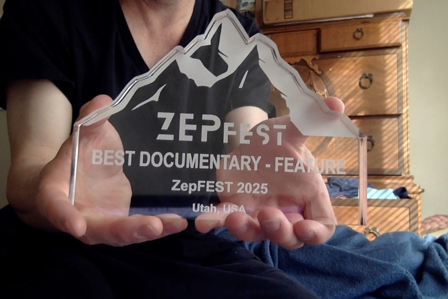 Person holding an acrylic award with mountain designs and text that reads 'ZepFEST BEST DOCUMENTARY - FEATURE ZepFEST 2025 Utah, USA'. The person is wearing a black shirt, seated in front of a wooden dresser with clothing items.