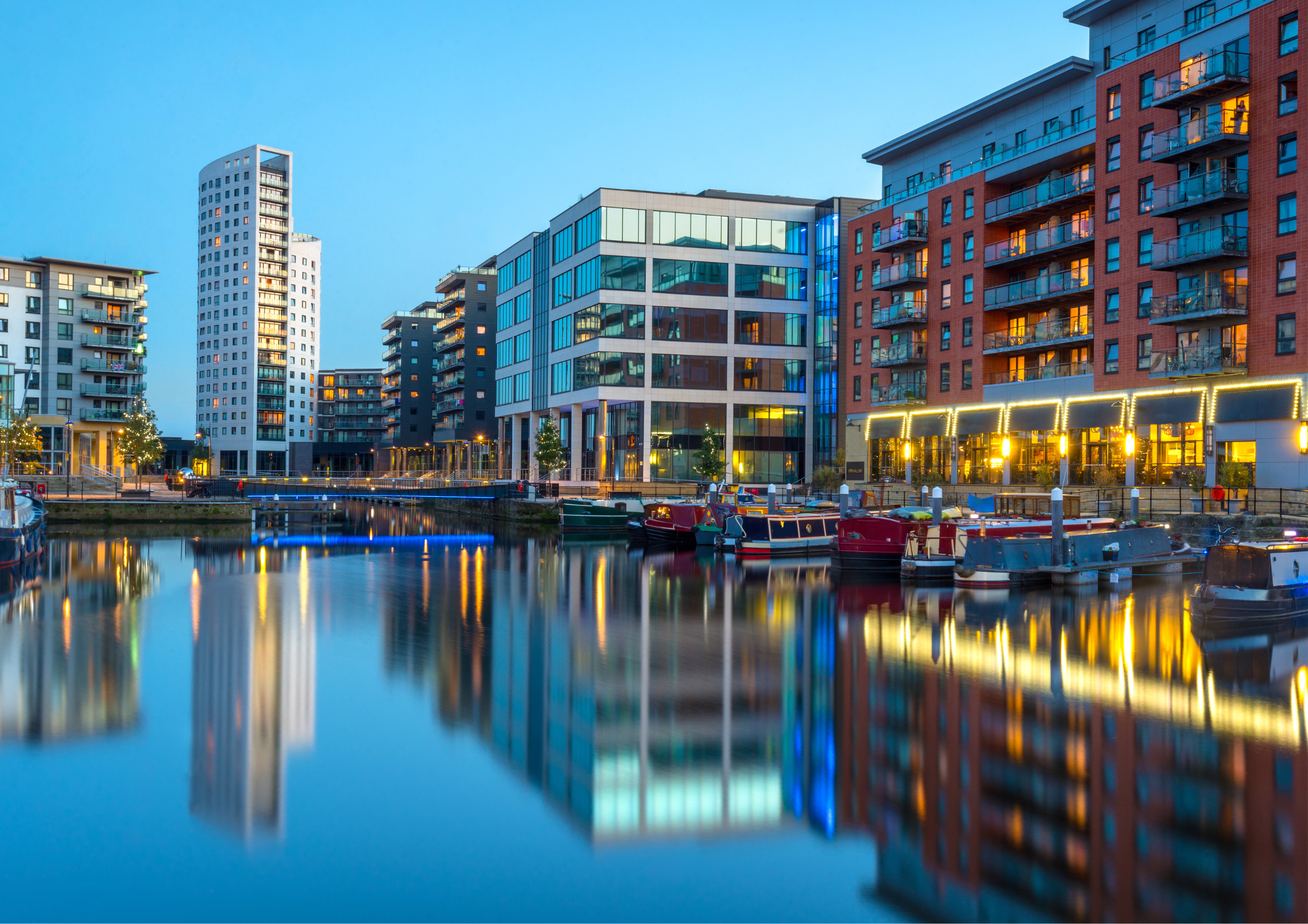Modern buildings along a waterfront with boats docked, during dusk with reflections in the water.