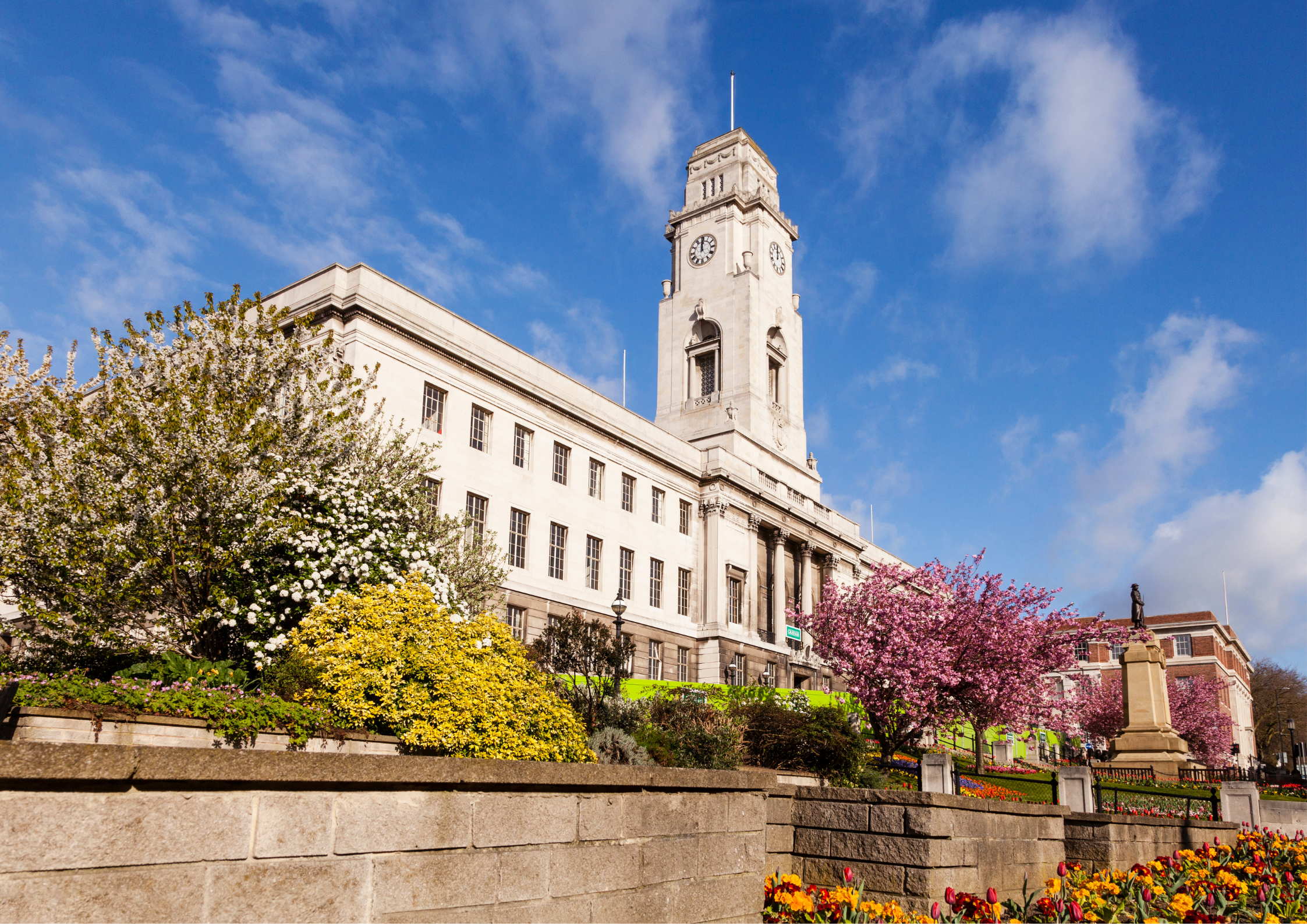 A historic building with a clock tower, surrounded by blooming trees and colorful flower beds, under a bright blue sky.