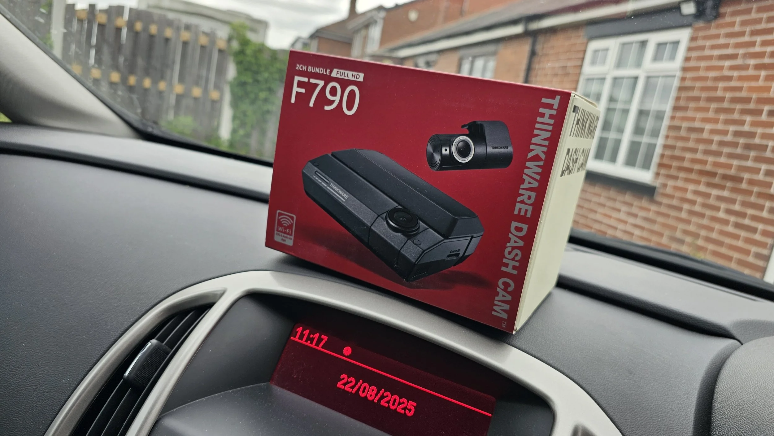 A boxed dash cam with a black camera and mount, labeled Thinnkware, sits on a car dashboard in front of a red digital clock display showing 11:17, inside a vehicle with a view of a residential brick house and fence outside the window.
