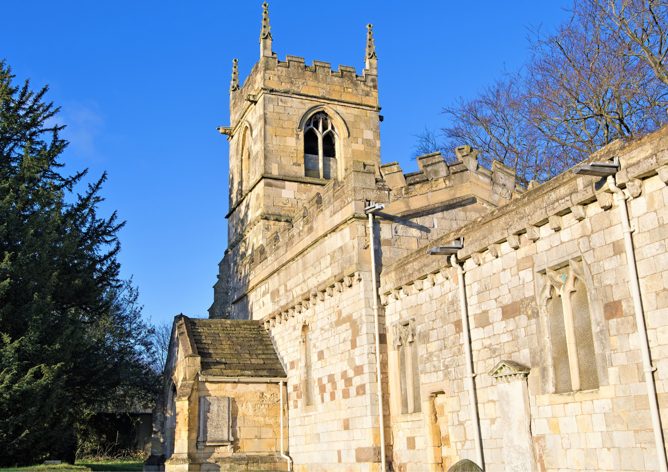 A stone church tower with gothic windows and decorative spires, part of an old church building, with trees and a clear blue sky in the background.