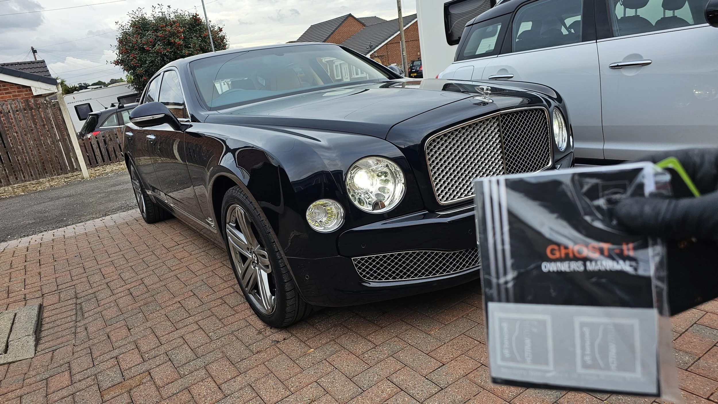 A black luxury car parked on a brick driveway next to a white vehicle. There is a fence with brick houses and a tree in the background, with cloudy sky overhead. A Ghost-11 owner's manual is partially visible in the foreground.
