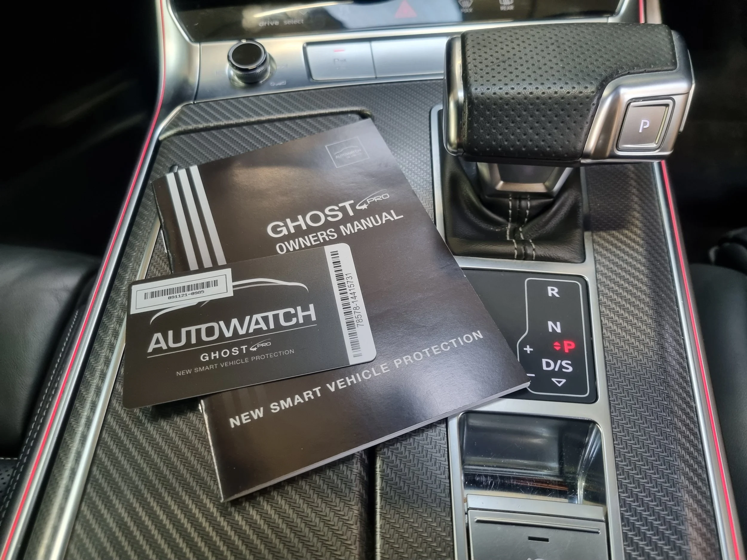 The image shows the center console of a car, with a Ghost AutoWatch owner's manual placed on the gear shift area. The gear shift is in the park position, and there is a carbon-fiber pattern surrounding the console.