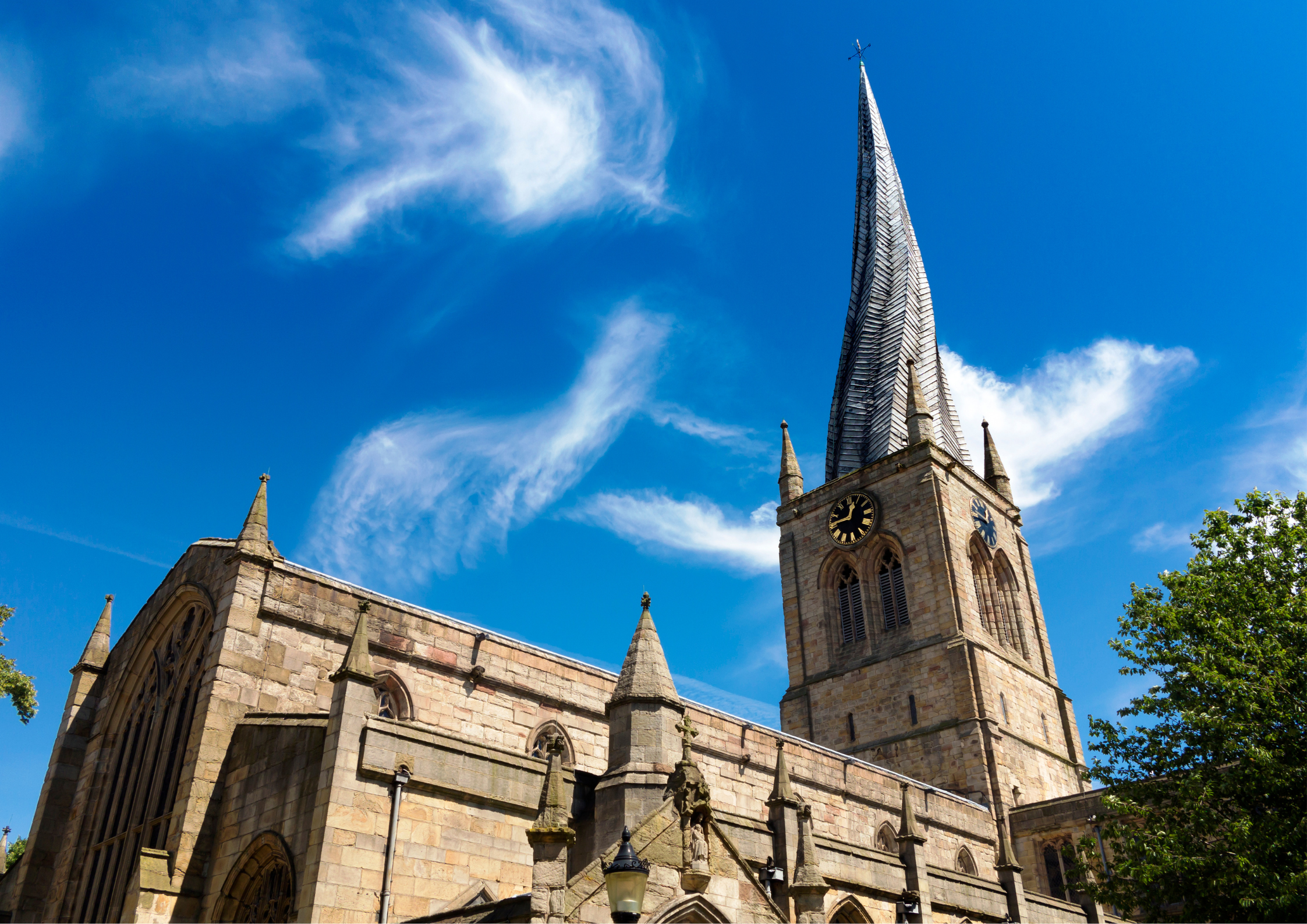 A stone church with a tall, pointed steeple against a bright blue sky with a few clouds. The church features Gothic-style architecture with arched windows and detailed stonework.