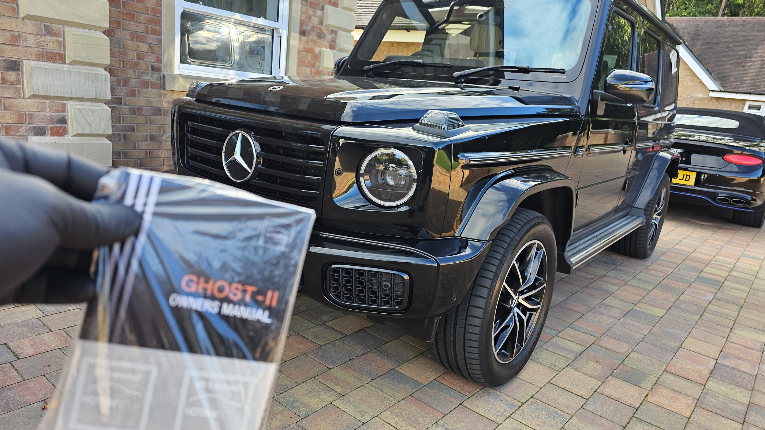Front view of a black Mercedes-Benz G-Class SUV parked outside a brick house, with a person holding a 'Ghost-II Owner's Manual' in the foreground.