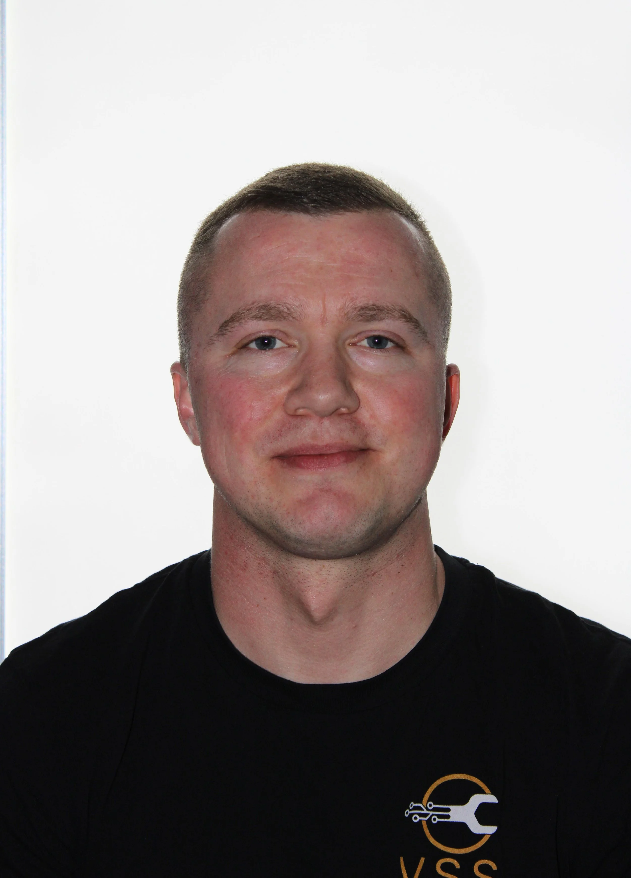 A young man with short brown hair, light skin, and blue eyes, wearing a black T-shirt with a logo on it, standing against a plain white background.