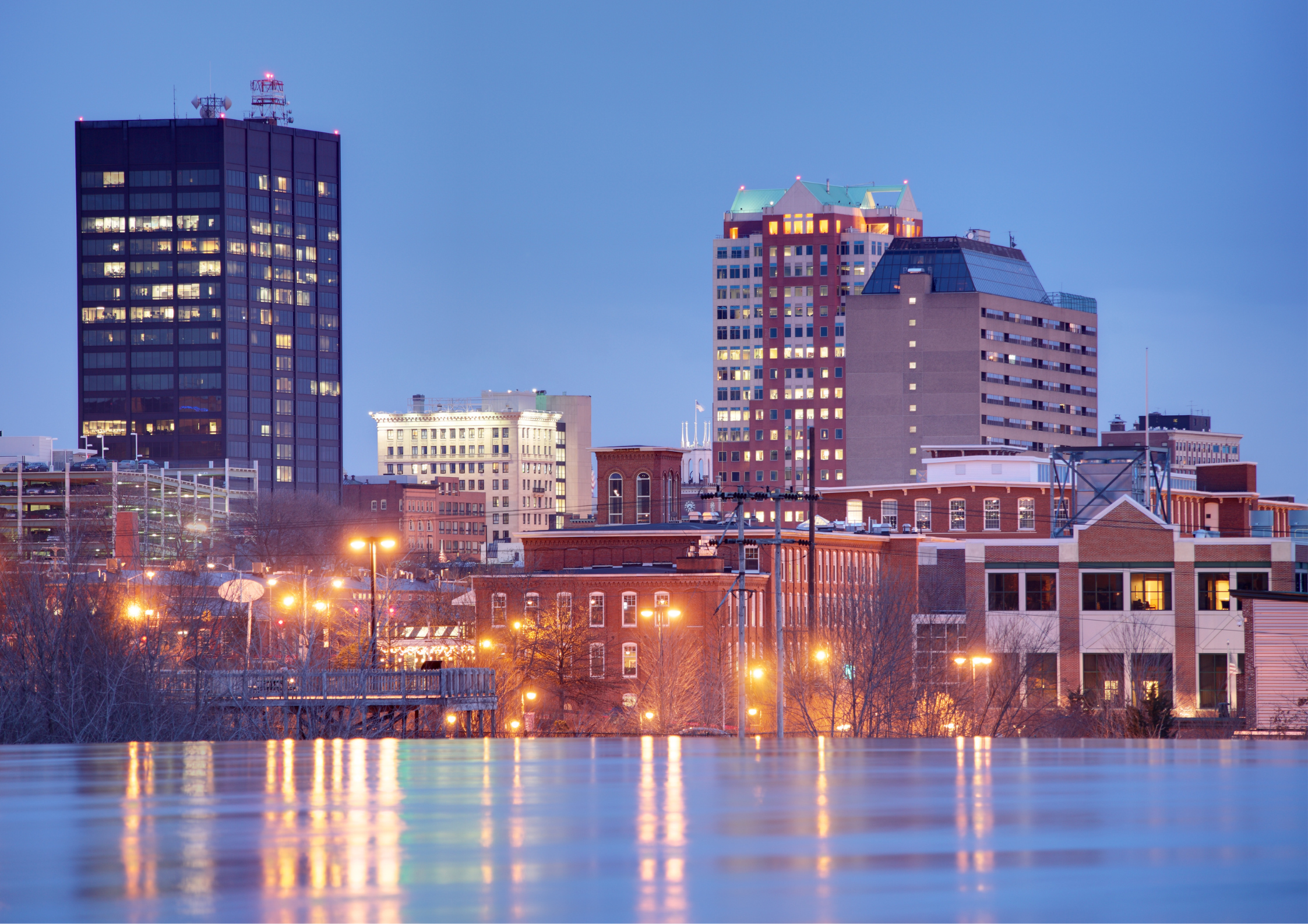 Nighttime city skyline with illuminated buildings and a body of water in the foreground.