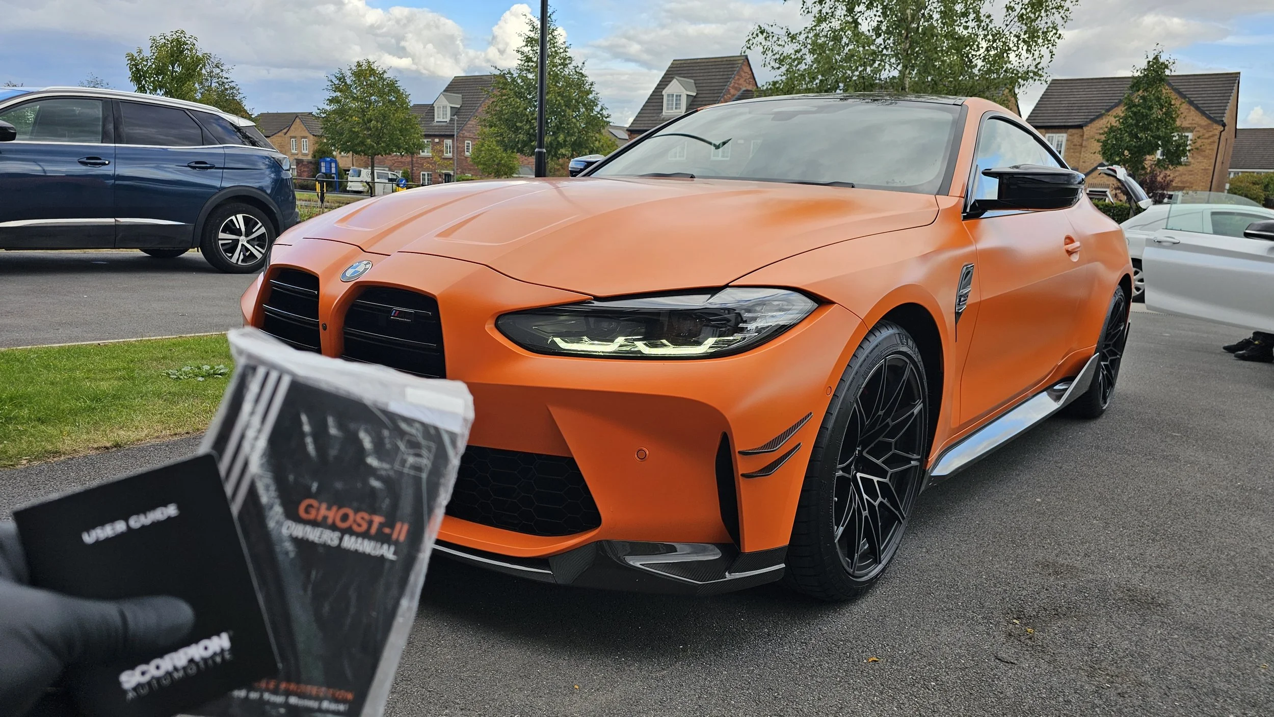 Orange BMW sports car parked in a lot, with a black garage door user guide in the foreground and other cars and houses in the background.