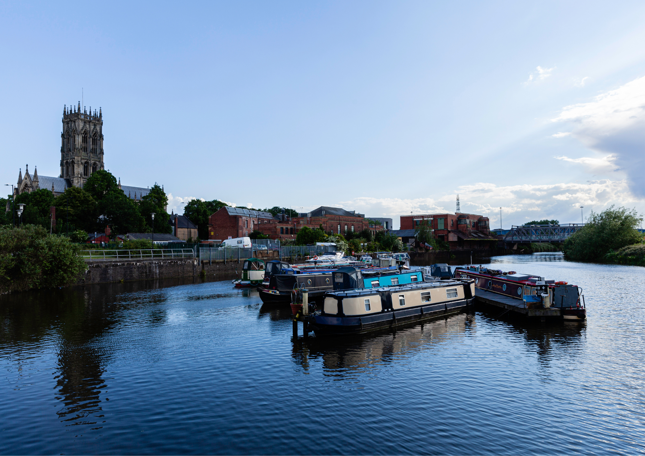 A view of a river with several boats docked along the edge, a historic church with a tall spire on the left, red brick buildings, and a bridge over the water. The sky is mostly clear with some clouds.