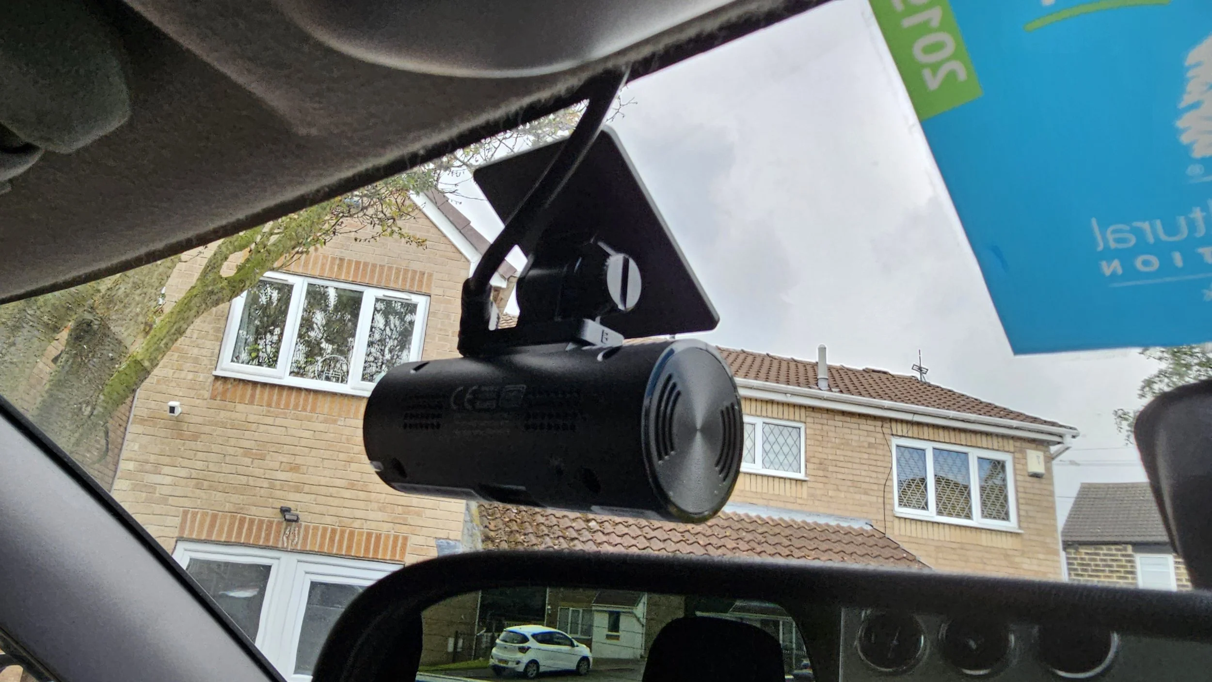 Photo taken from inside a car, showing the dashboard and rearview mirror. The mirror has a dash camera attached. In the background, residential houses with brick walls and tiled roofs, a tree, and a parked white car are visible. Part of a blue and green sign is also seen.