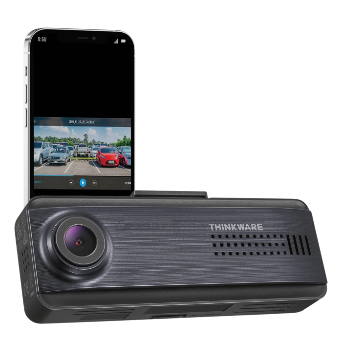 A Thinkware dash cam with a smartphone showing a parking lot with parked cars on its screen.