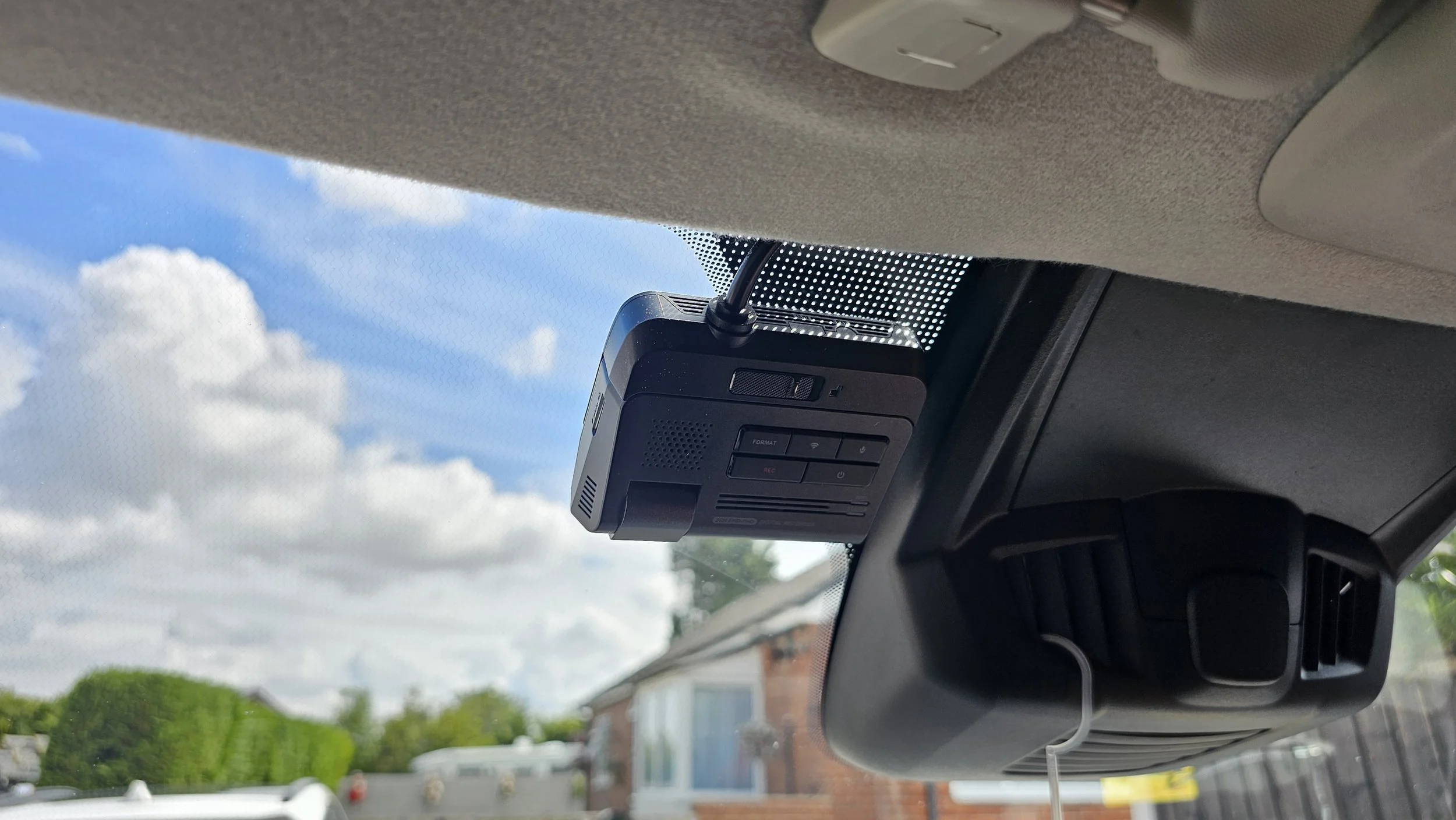In car interior, dashboard camera mounted on the ceiling, visible in front of the windshield with sky and clouds outside.