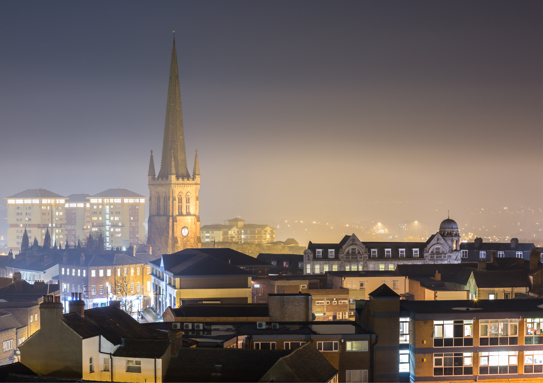 Nighttime cityscape with illuminated buildings, a church with a tall spire, and a dark sky with fog or haze.