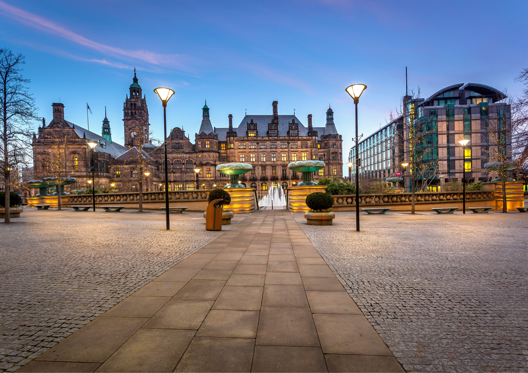 A city square with a historic building featuring towers, and a modern glass building, decorated with string lights on trees, during evening twilight.