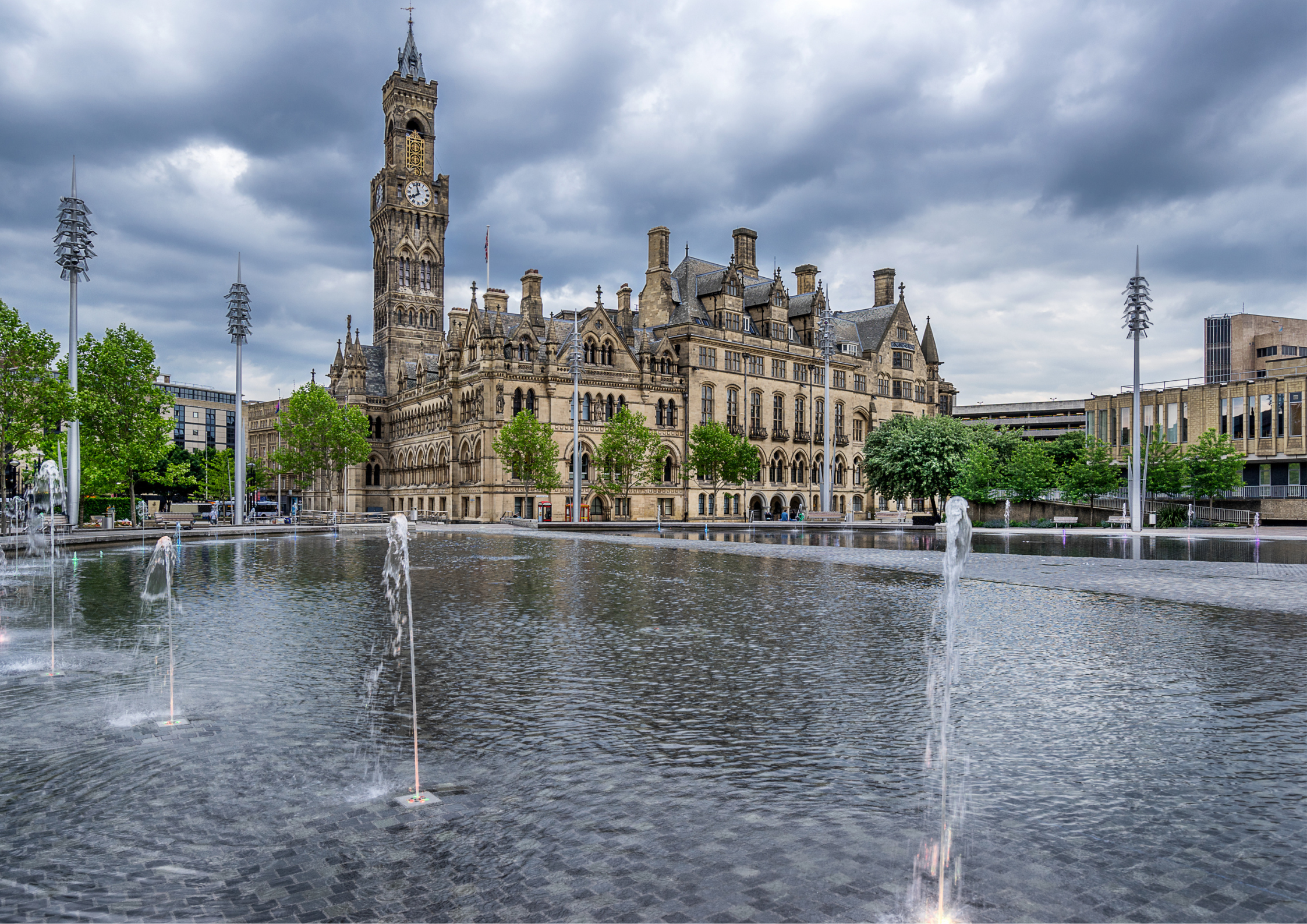 A historic building with a tall clock tower and Gothic architectural details, viewed across a fountain in a park with trees and modern buildings in the background.