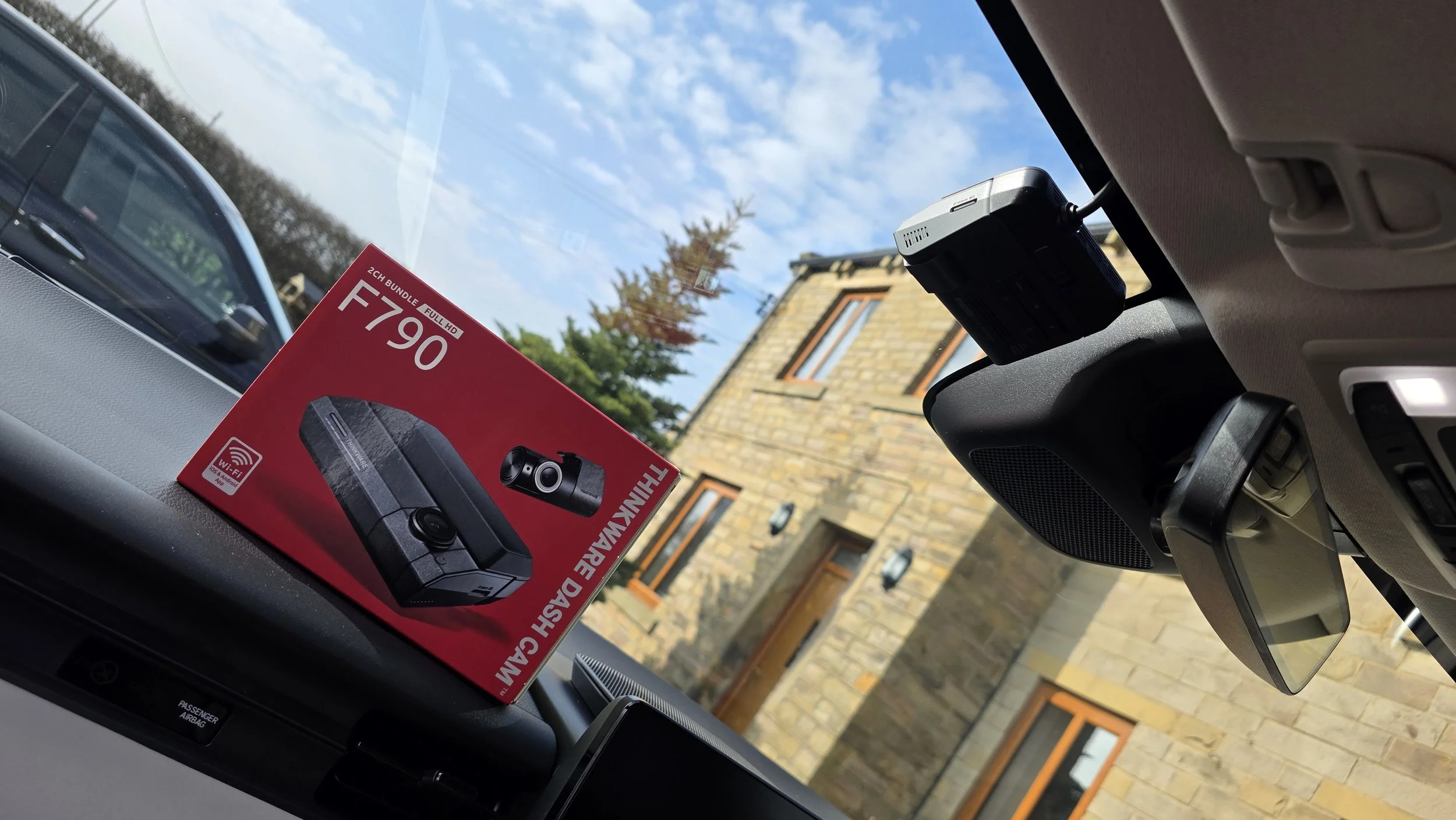 Car interior showing a dashboard with a Thule box set on the windshield and a rearview mirror. Outside view shows a building with stone walls and trees under a partly cloudy sky.