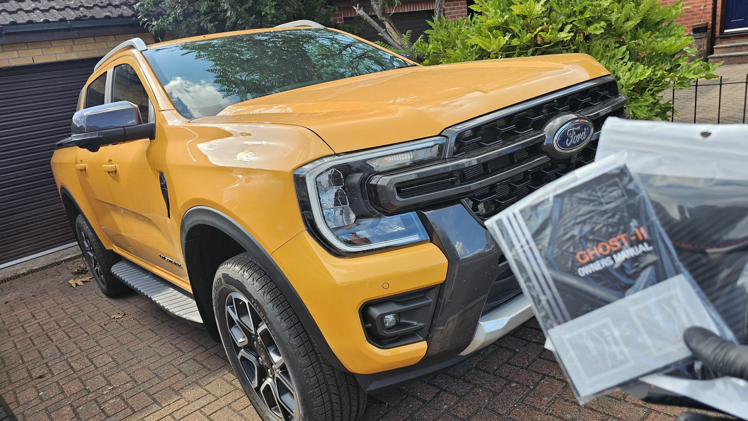 A yellow Ford pickup truck parked on a brick driveway, with lush green bushes and a brick house in the background. A person is holding a clear plastic bag with a Ghost-II owner's manual in the foreground.