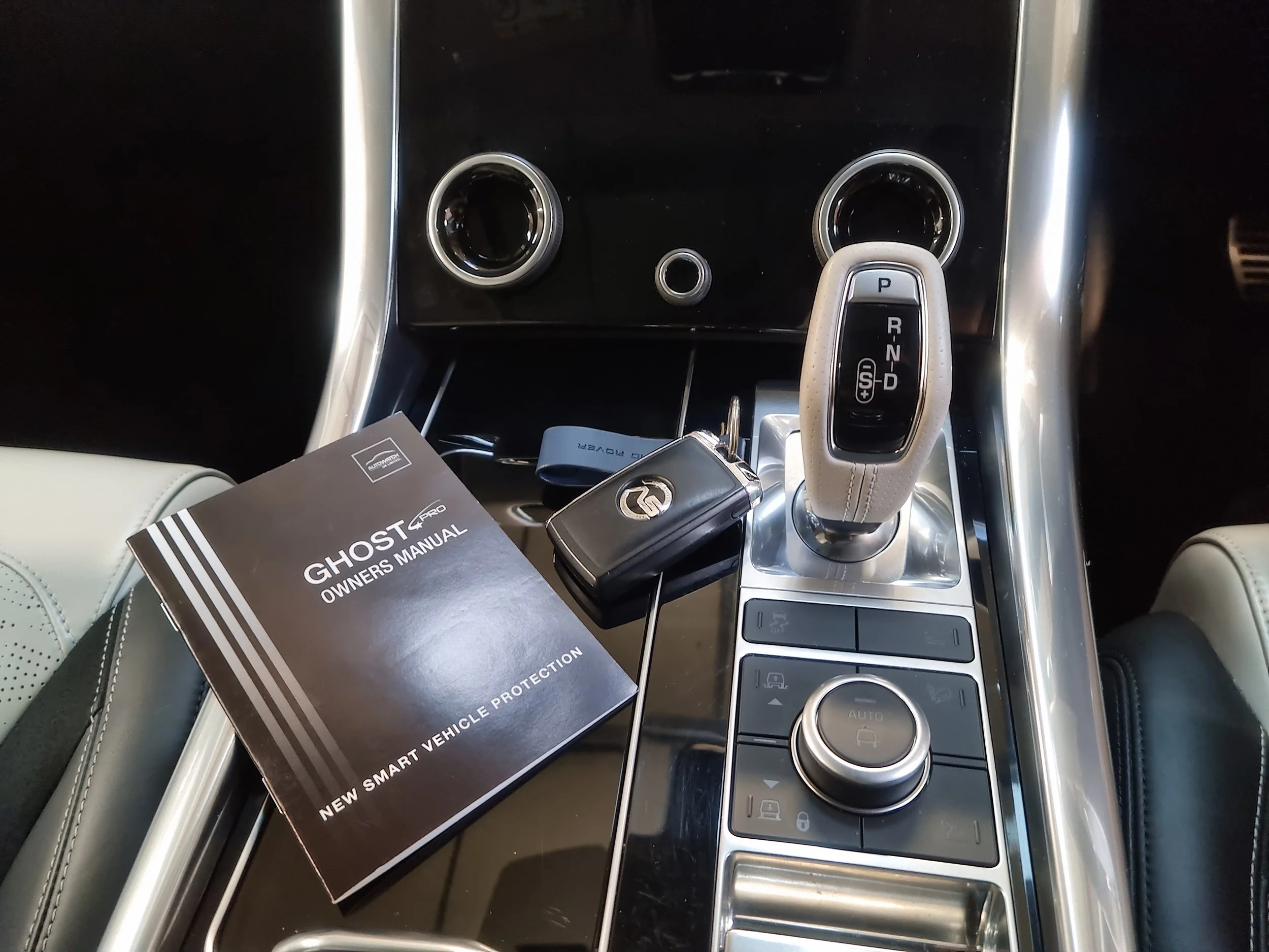 Close-up view of a car's center console showing an automatic gear shift lever, two cup holders, a small circular port, and a manual car key with a black cover placed on the console. There is also a booklet titled 'GHOST PRO' partially visible beneath the key.