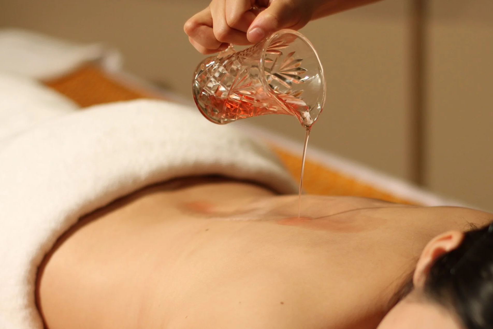 Warm massage oil in a glass jar is poured down the back of a massage client.