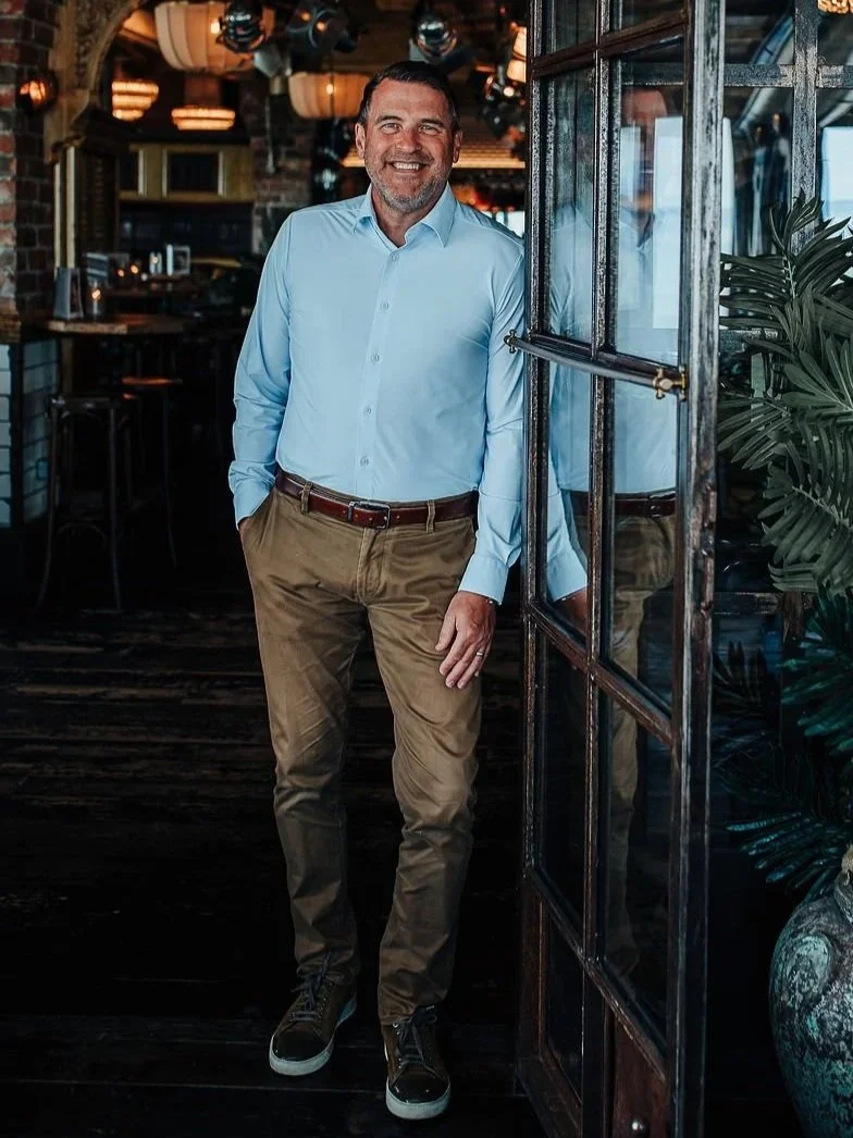 A smiling man in a light blue dress shirt and khaki pants standing by an open glass door in a restaurant or cafe.