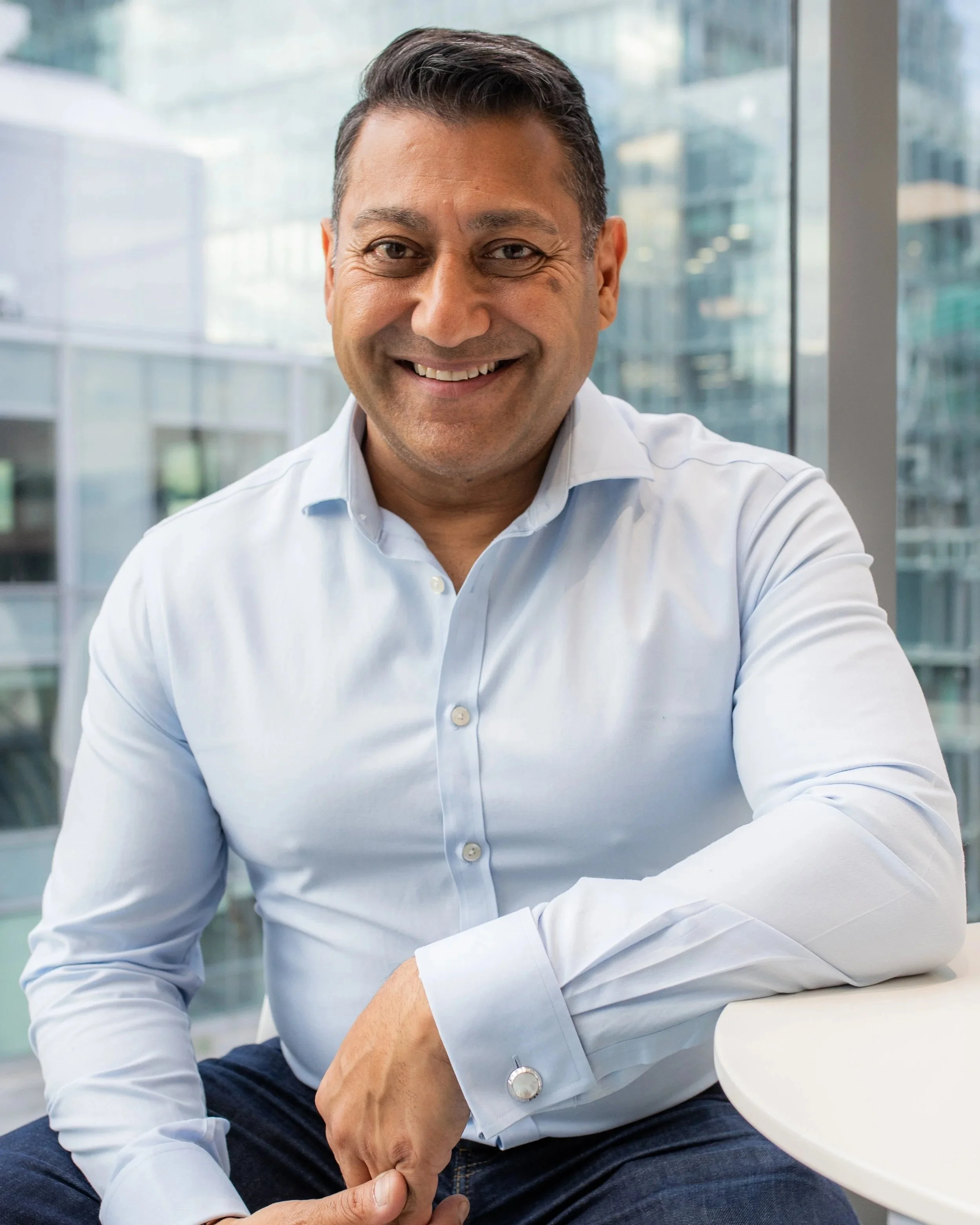 Smiling man in a white shirt sitting indoors near large windows with a cityscape in the background.