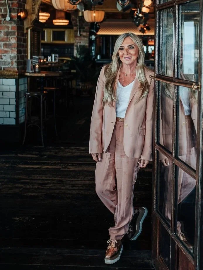 A woman in a beige suit standing inside a restaurant with brick walls and warm lighting.