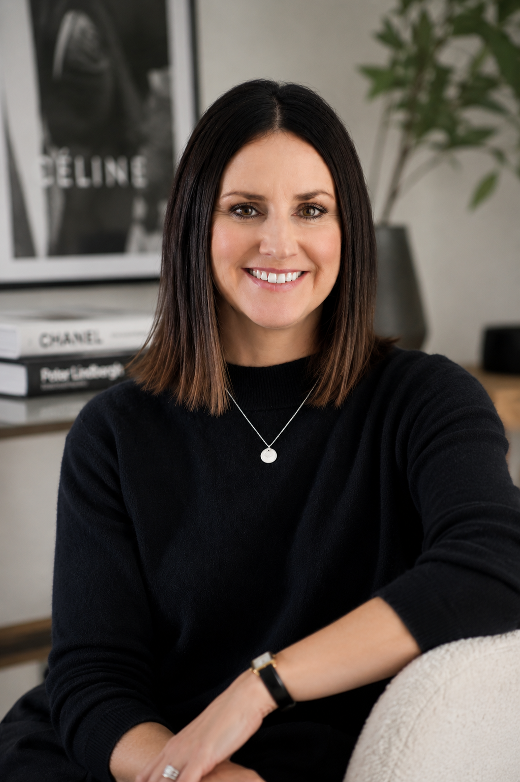 A woman with shoulder-length dark brown hair, wearing a black sweater, silver necklace, watch, and rings, smiling while sitting on a cream-colored chair in a modern living room with plants and art in the background.