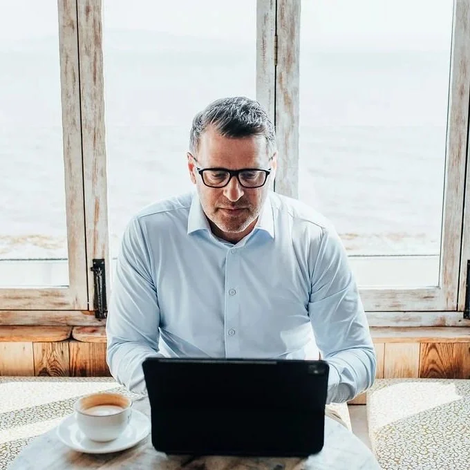 Man wearing glasses and a light blue shirt working on a laptop at a table with a cup of coffee, near large windows with a view of water.