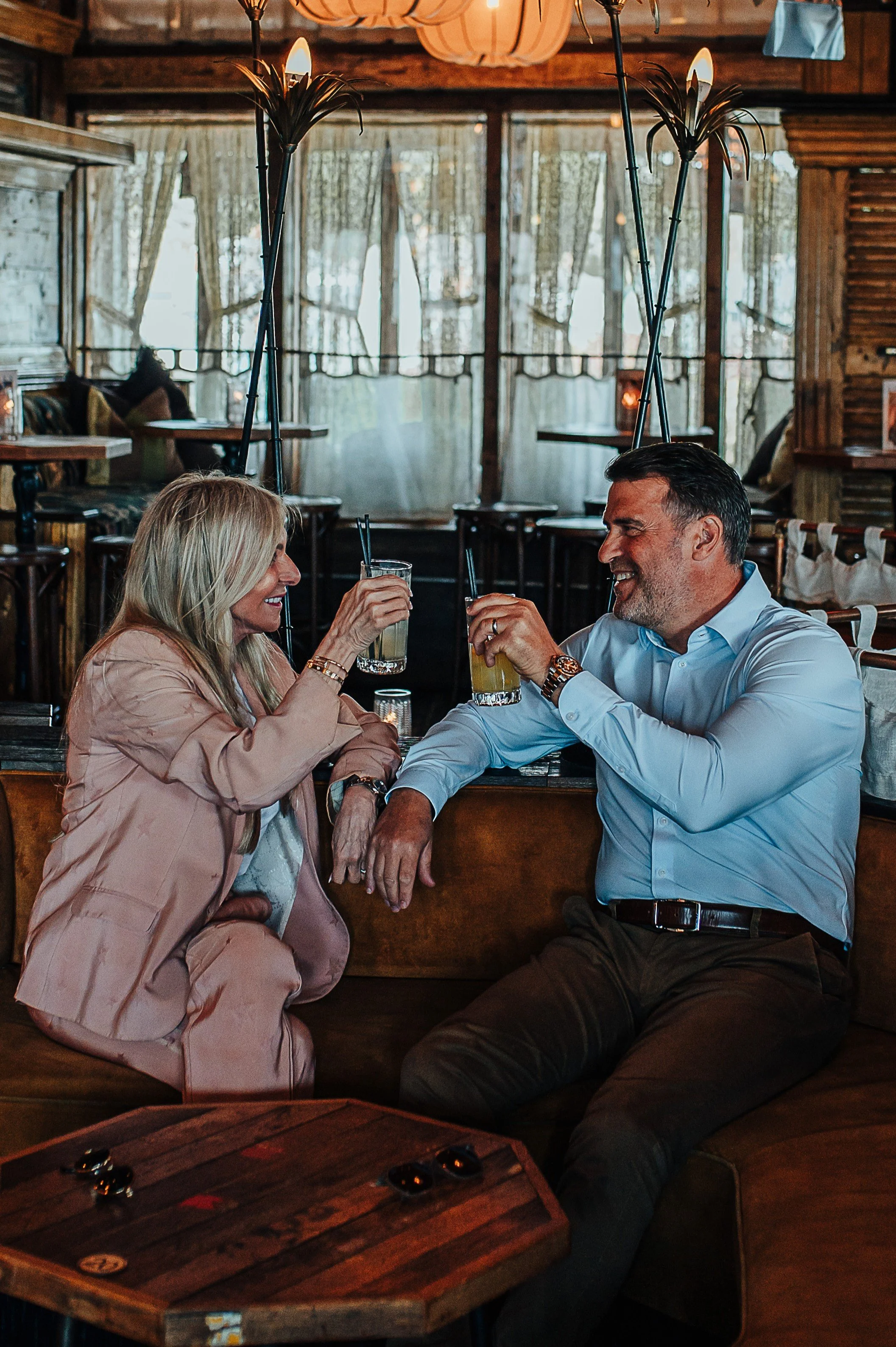 A middle-aged man and woman are sitting at a cozy wooden table in a rustic restaurant, sharing drinks and smiling at each other. The woman is wearing a light pink blazer and has long blonde hair, while the man is in a light blue dress shirt with rolled-up sleeves, and dark pants. Behind them are large windows with curtains, and the room is decorated with wooden walls and warm lighting.