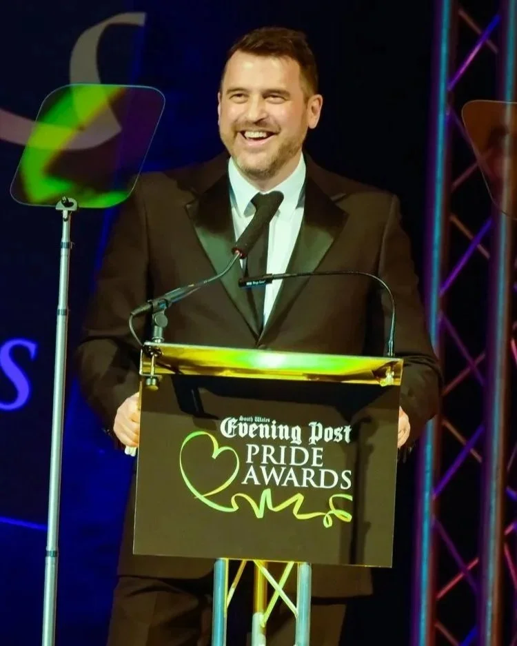 A man in a tuxedo standing at a podium with a sign that reads 'South Alabama Evening Post Pride Awards.' He is smiling and speaking into a microphone, with stage lighting and decorations in the background.