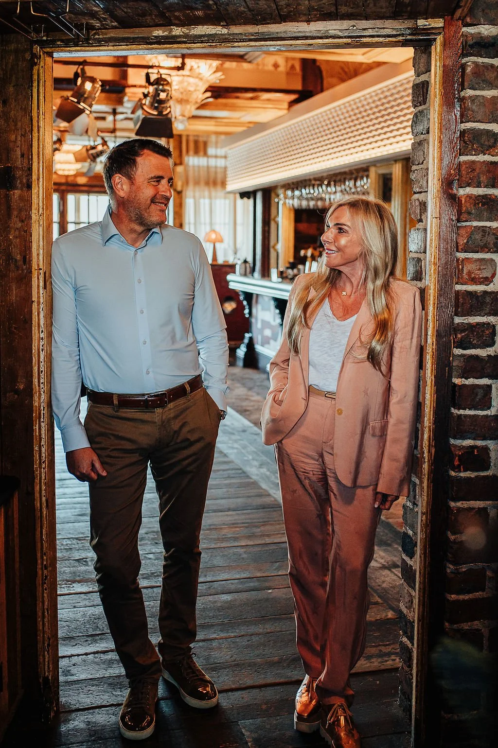 A man and woman are talking and smiling in a rustic, warmly lit restaurant with wood accents and a bar in the background.
