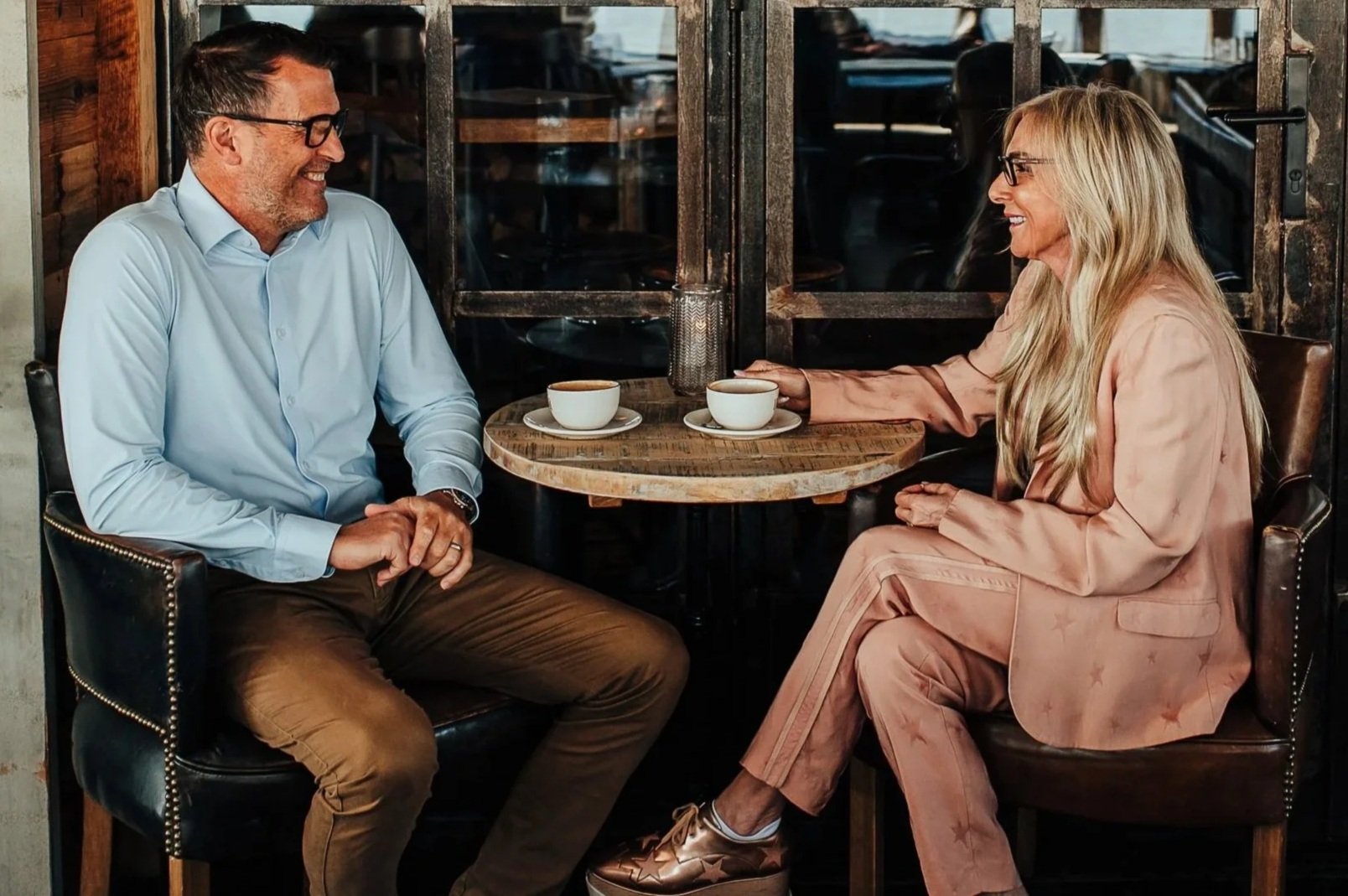 A man and a woman sit at a small round wooden table in a cafe, smiling and enjoying coffee together. They are facing each other and engaged in a pleasant conversation.