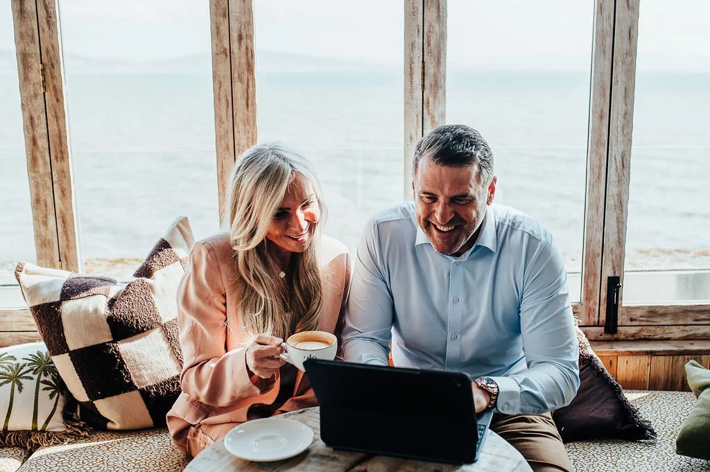 A man and woman sitting together indoors, smiling at a laptop on the table, with the woman holding a cup, and a window with a view of the beach outside.